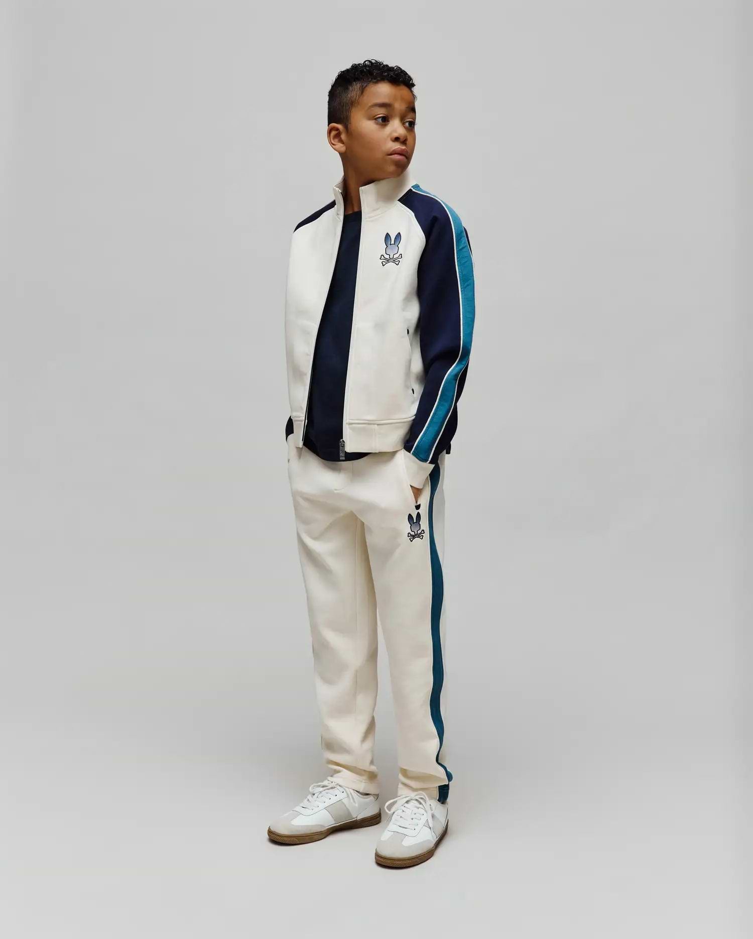 Cream track suit: jacket with navy/teal color block sleeves, chest Psycho Bunny skull logo; KIDS Caspian Contrast Stripe Track Pant with teal side stripes, thigh bunny skull logo. Classic fit.