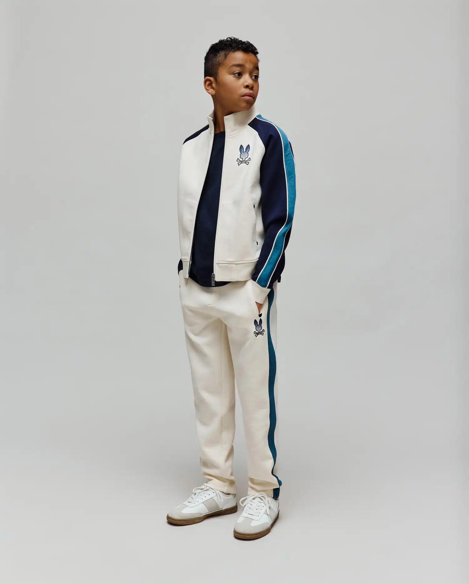 Cream track suit: jacket with navy/teal color block sleeves, chest Psycho Bunny skull logo; KIDS Caspian Contrast Stripe Track Pant with teal side stripes, thigh bunny skull logo. Classic fit.