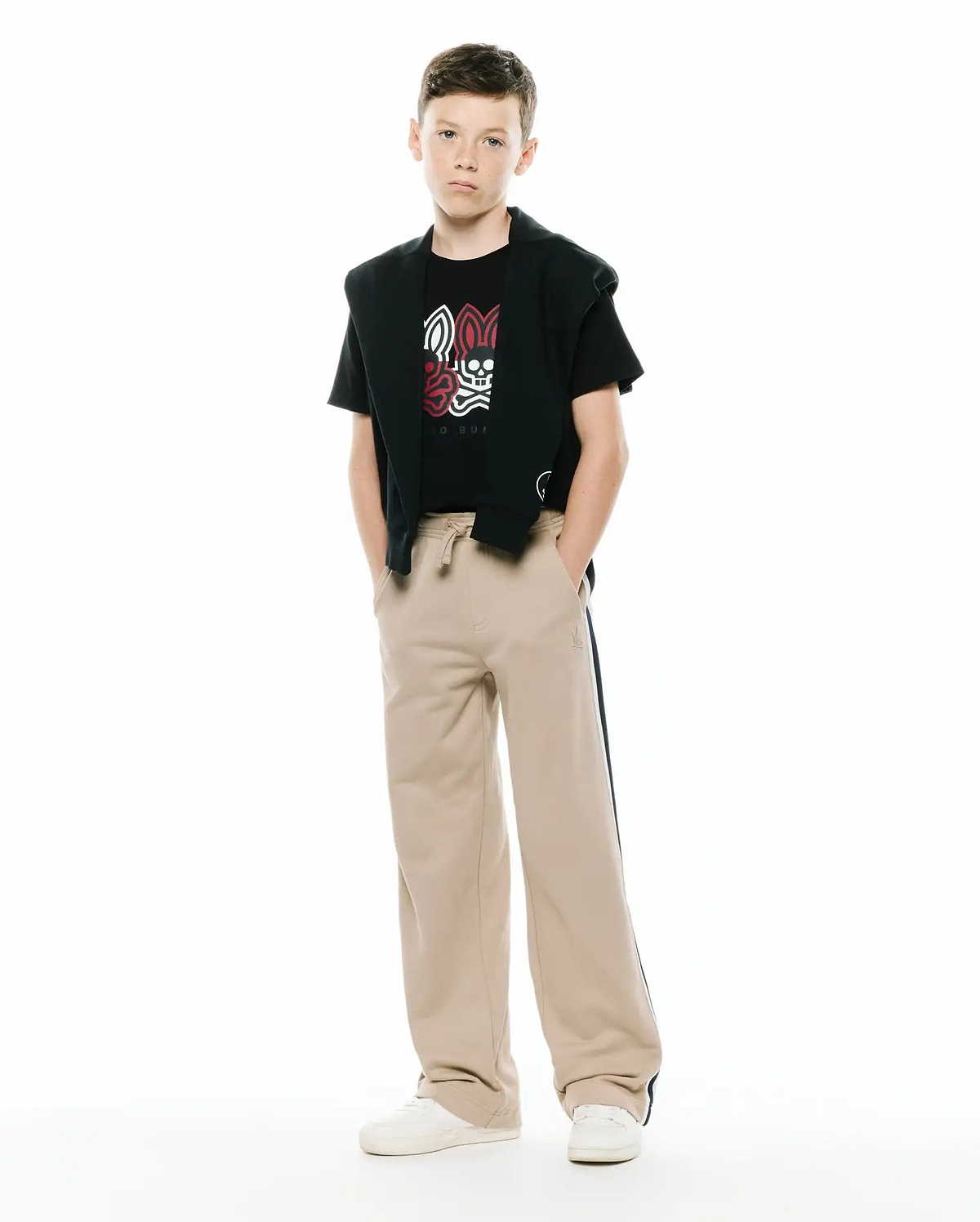 Black jersey t-shirt with a large white and red Psycho Bunny skull graphic, paired with a black French terry hoodie draped over shoulders and KIDS TY FRENCH TERRY SWEATPANT in beige with a dark side stripe and small embroidered logo.