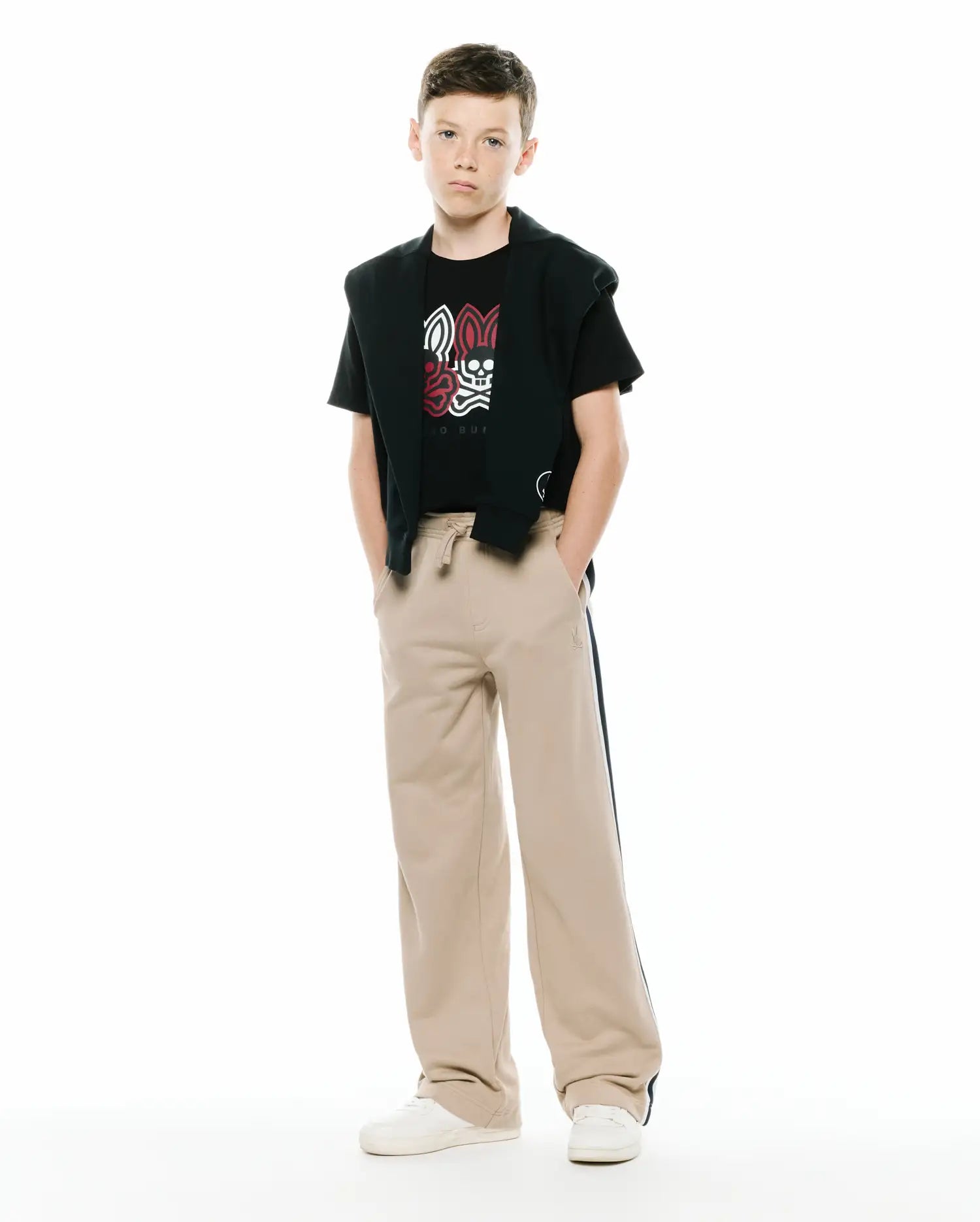 Black jersey t-shirt with a large white and red Psycho Bunny skull graphic, paired with a black French terry hoodie draped over shoulders and KIDS TY FRENCH TERRY SWEATPANT in beige with a dark side stripe and small embroidered logo.