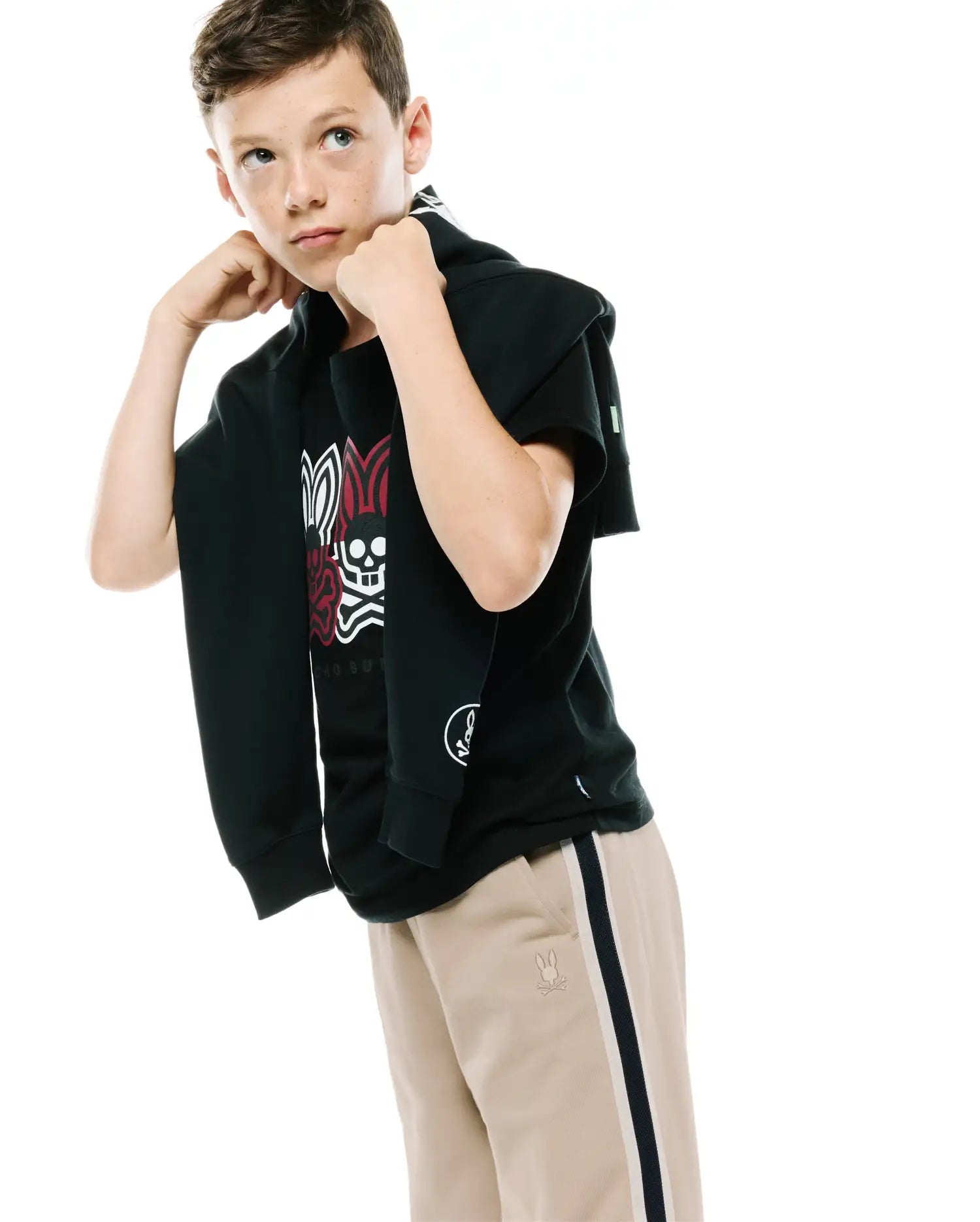 Black jersey t-shirt with large red, white, and black Psycho Bunny skull graphic on back, paired with Kids Ty French Terry Sweatpant in khaki featuring a dark blue and white side stripe and embroidered Psycho Bunny logo on the left thigh.