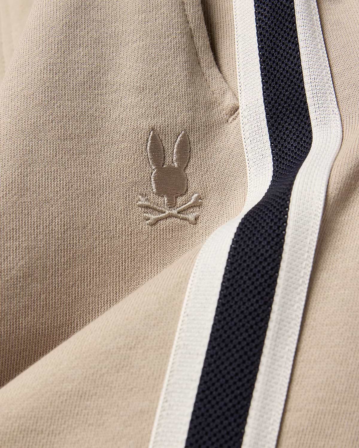Kids Ty French Terry Sweatpant in beige french terry fabric, featuring a small embroidered Psycho Bunny skull logo and a navy and white side stripe.