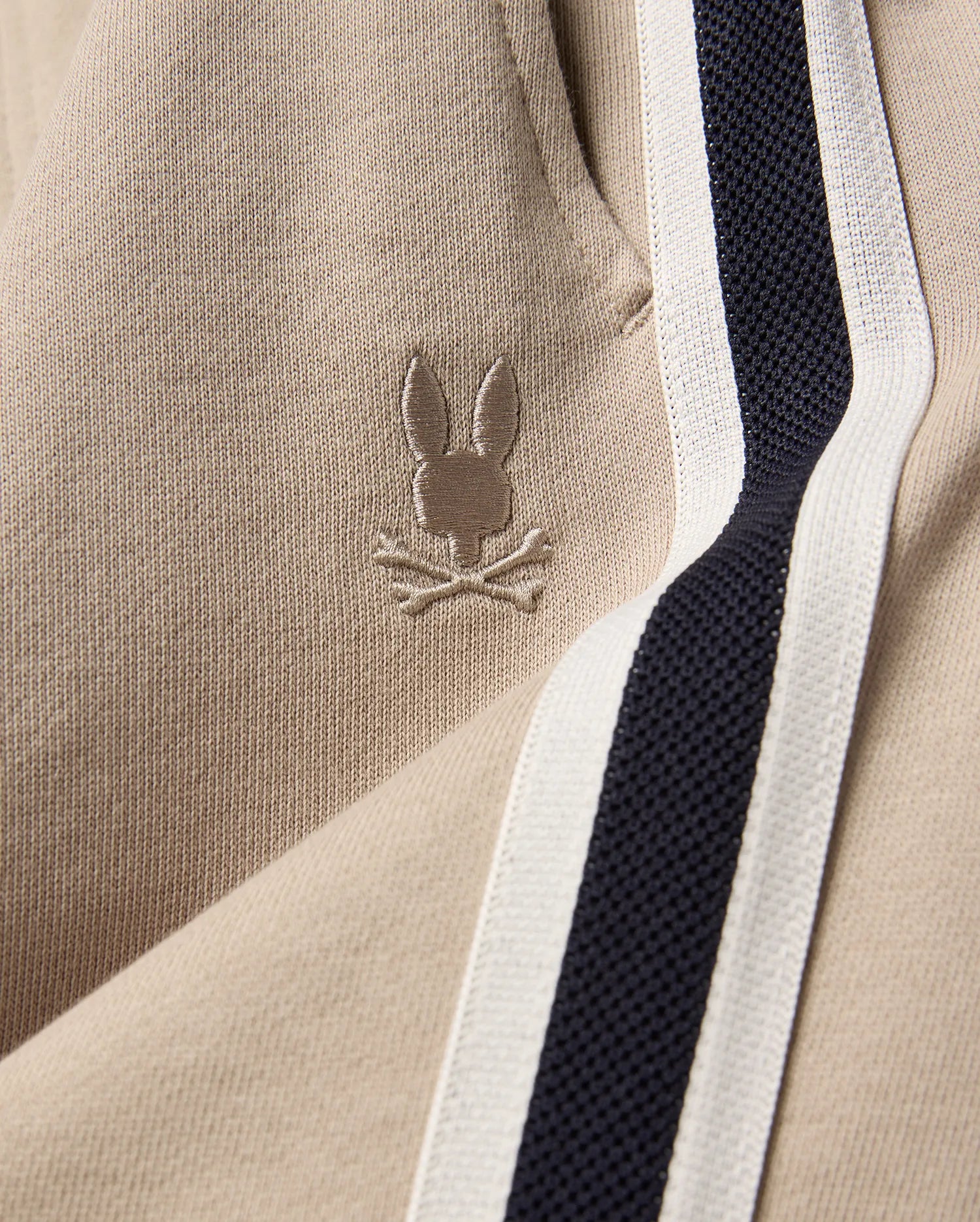 Kids Ty French Terry Sweatpant in beige french terry fabric, featuring a small embroidered Psycho Bunny skull logo and a navy and white side stripe.