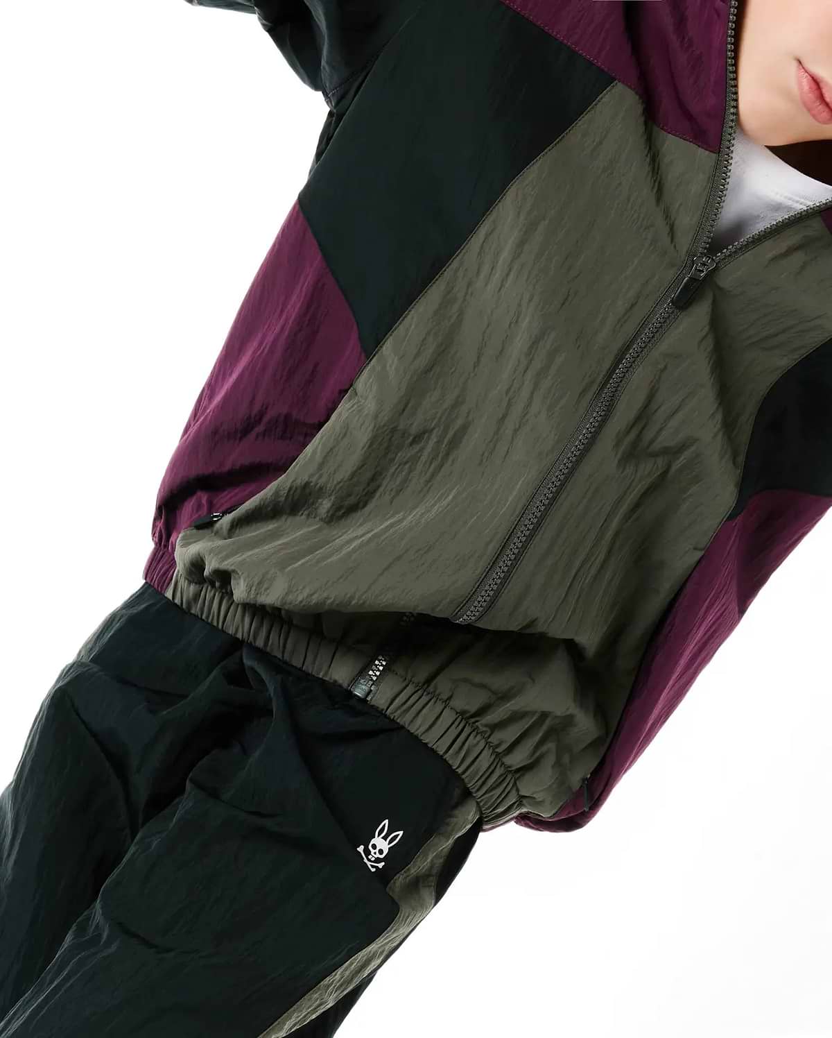 Kids Alder Nylon Colorblock Pant and matching jacket in olive green, black, and deep purple, featuring a relaxed fit, crinkled nylon fabric, and a small white embroidered Psycho Bunny skull logo on the left thigh.