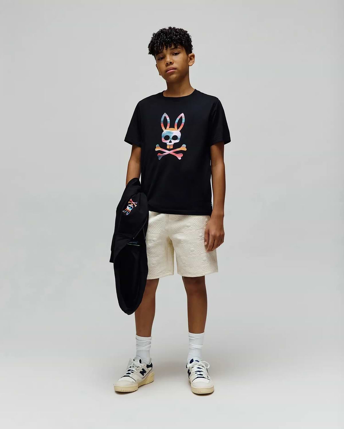 Black jersey t-shirt with a large multicolor Psycho Bunny skull graphic, paired with cream KIDS DUSTIN SWEATSHORT featuring an allover textured pattern, and a black French terry hoodie with a small embroidered logo.