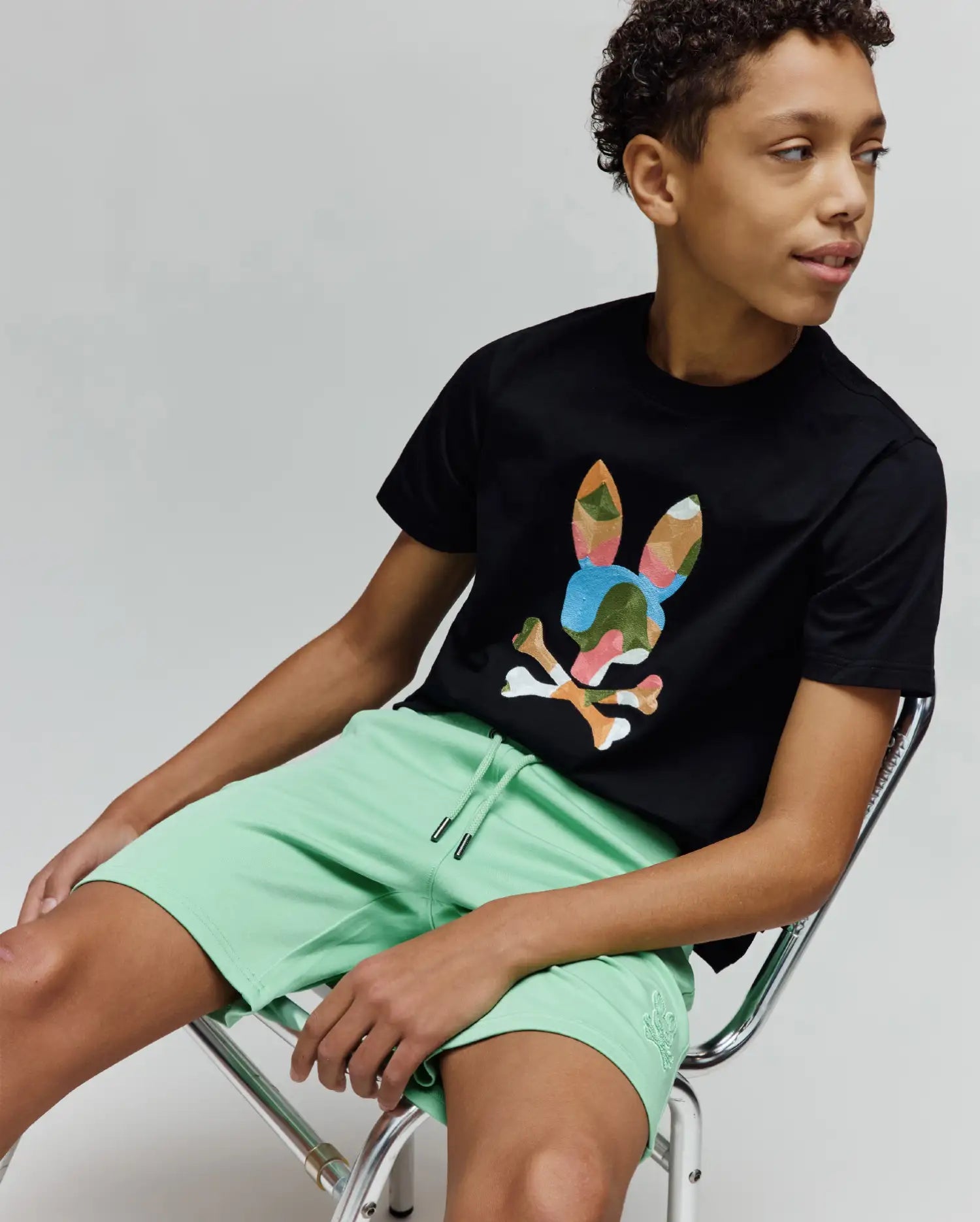 Black jersey t-shirt with a large multi-color camo Psycho Bunny skull graphic on the chest, paired with Kids Barry Modal Short in mint green featuring a drawstring waist and small tonal embroidered logo.