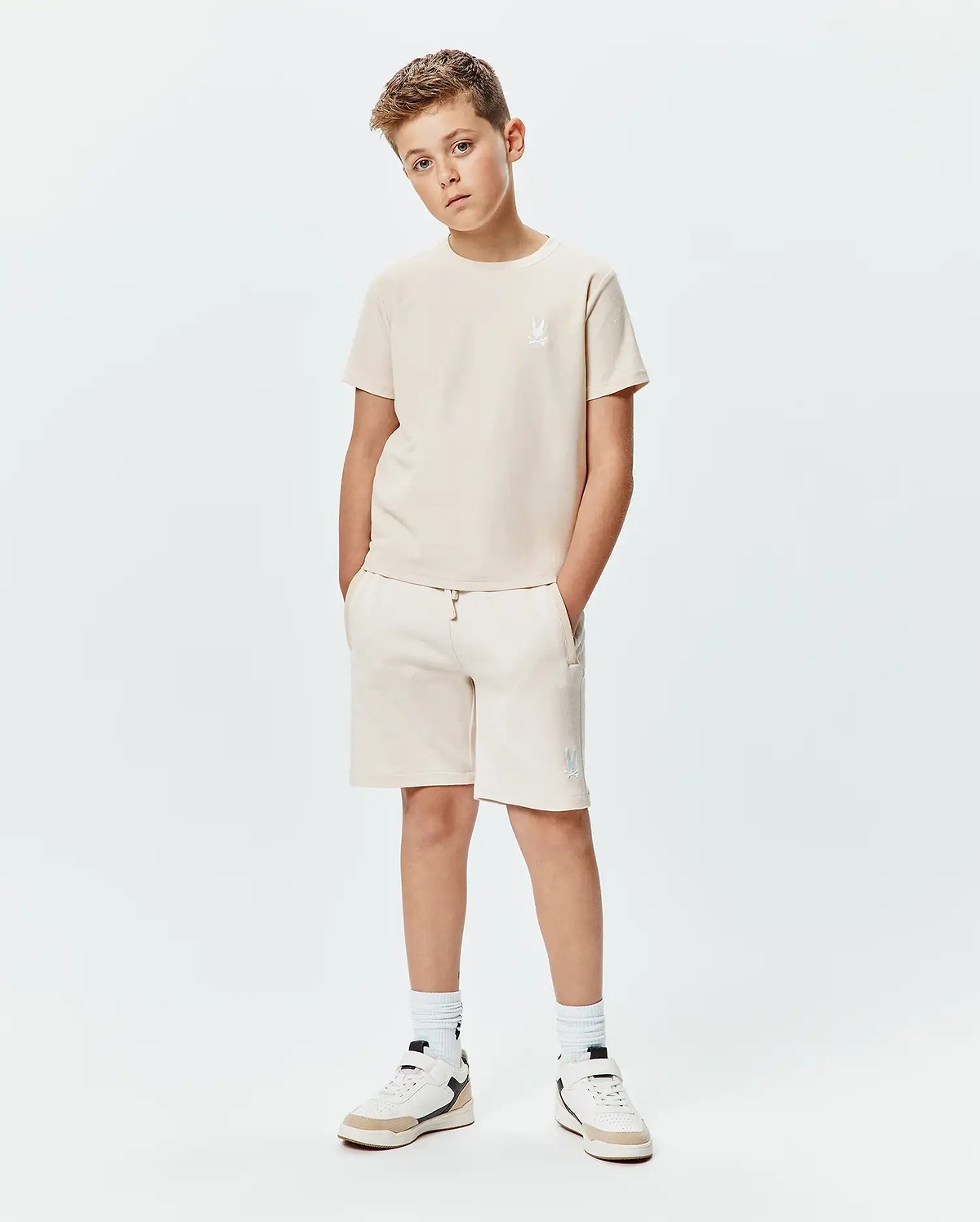 Psycho Bunny kids beige jersey t-shirt and Kids Keith Short 3 1/2 French terry shorts. Both feature white embroidered bunny skull logos, on the left chest and left thigh.