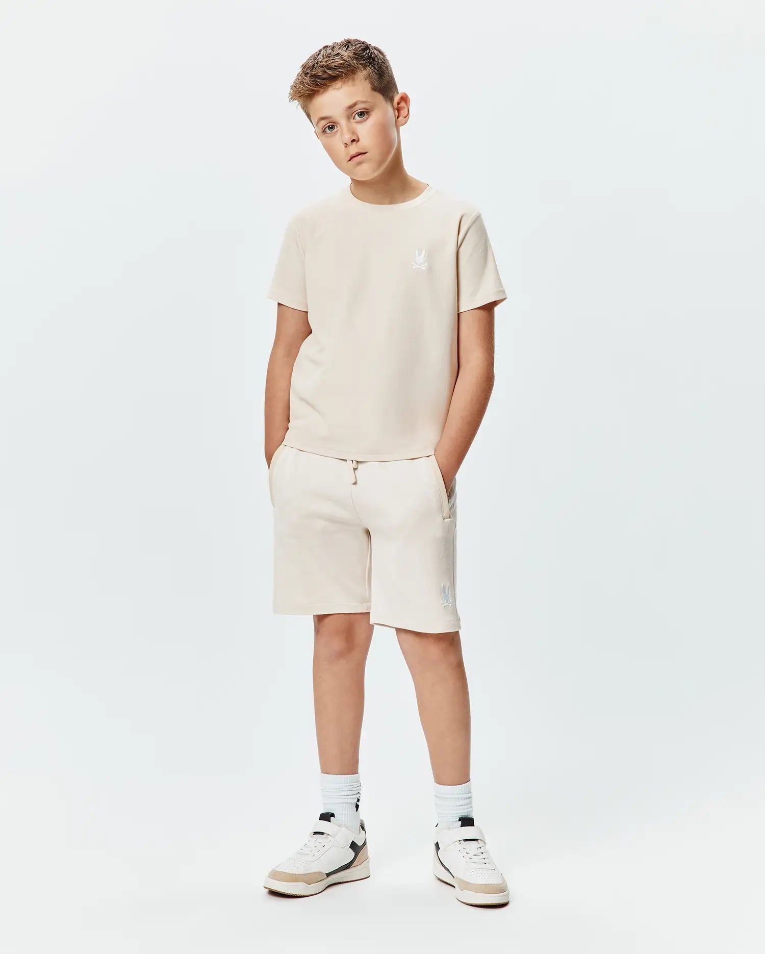 Psycho Bunny kids beige jersey t-shirt and Kids Keith Short 3 1/2 French terry shorts. Both feature white embroidered bunny skull logos, on the left chest and left thigh.