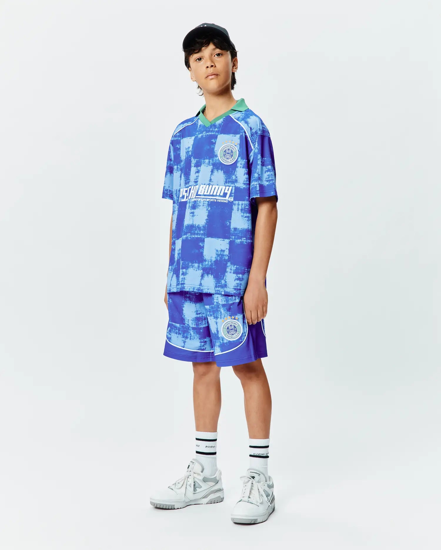 Psycho Bunny Kids Micah Soccer Short and matching relaxed-fit jersey polo in blue distressed checkered print, featuring a green collar, white piping, and circular Football Club logos on chest and leg.