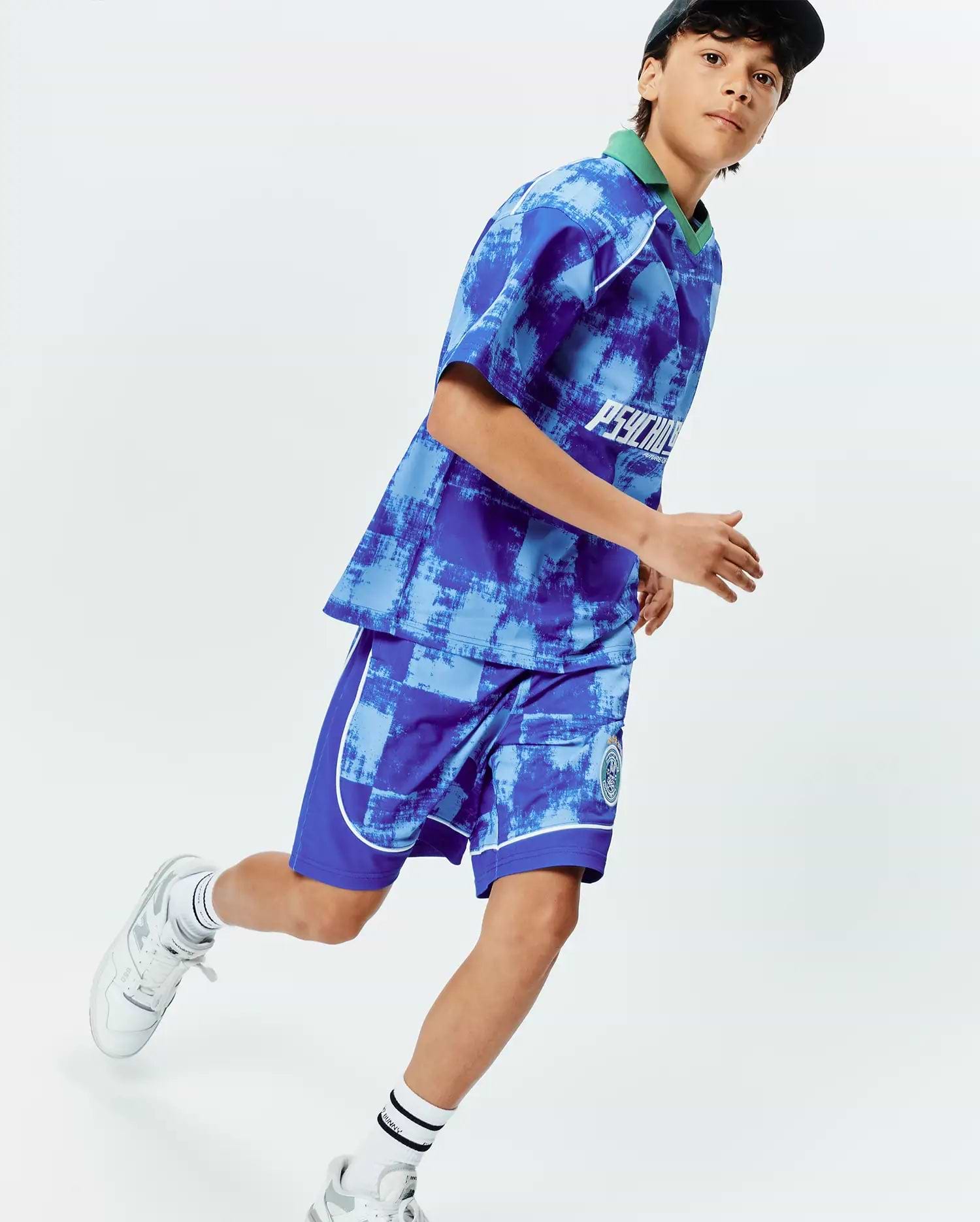 Vibrant blue and light blue distressed pattern jersey top with green collar, white Psycho Bunny chest text, and matching KIDS MICAH SOCCER SHORT with white piping and green bunny skull leg logo.