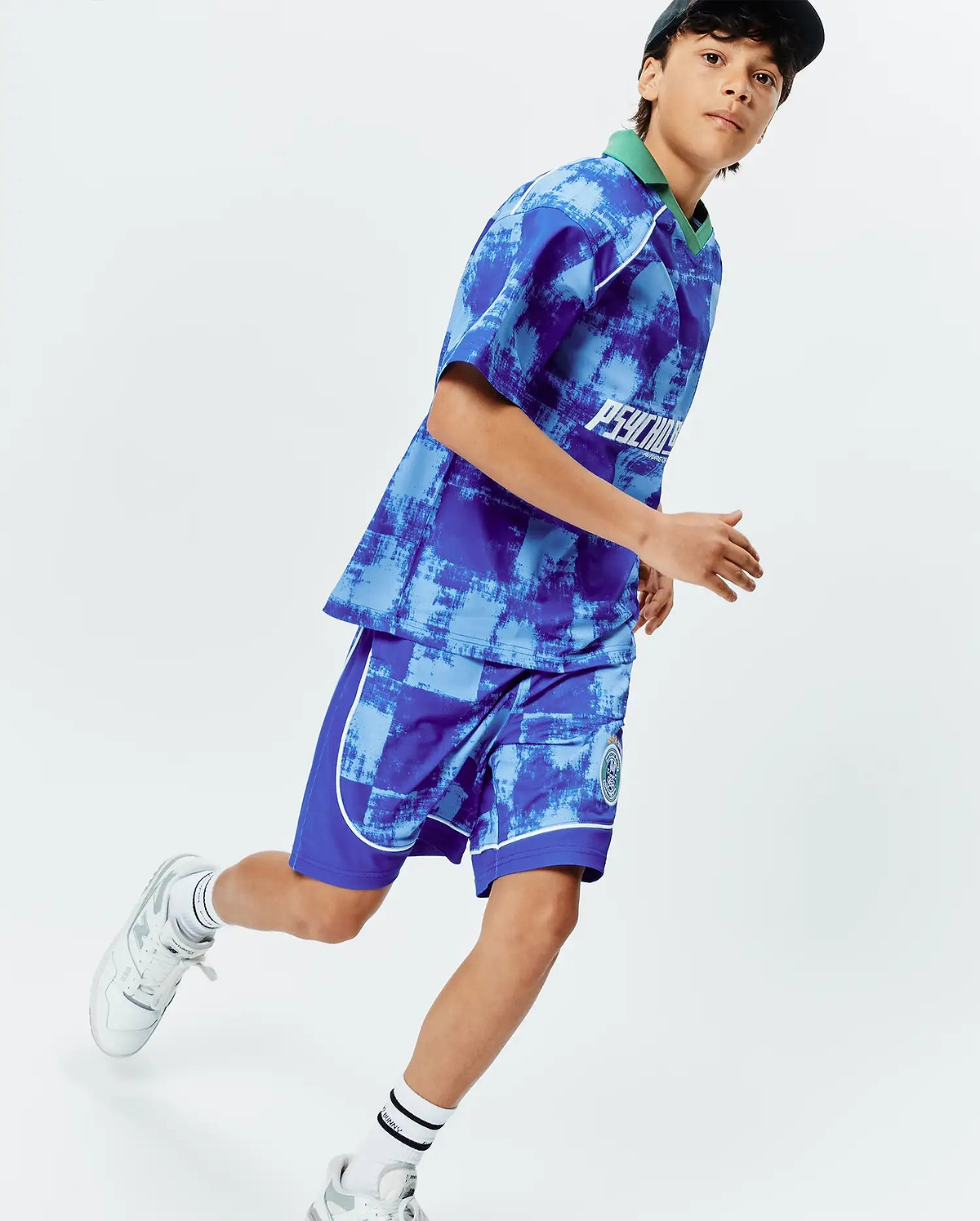 Vibrant blue and light blue distressed pattern jersey top with green collar, white Psycho Bunny chest text, and matching KIDS MICAH SOCCER SHORT with white piping and green bunny skull leg logo.
