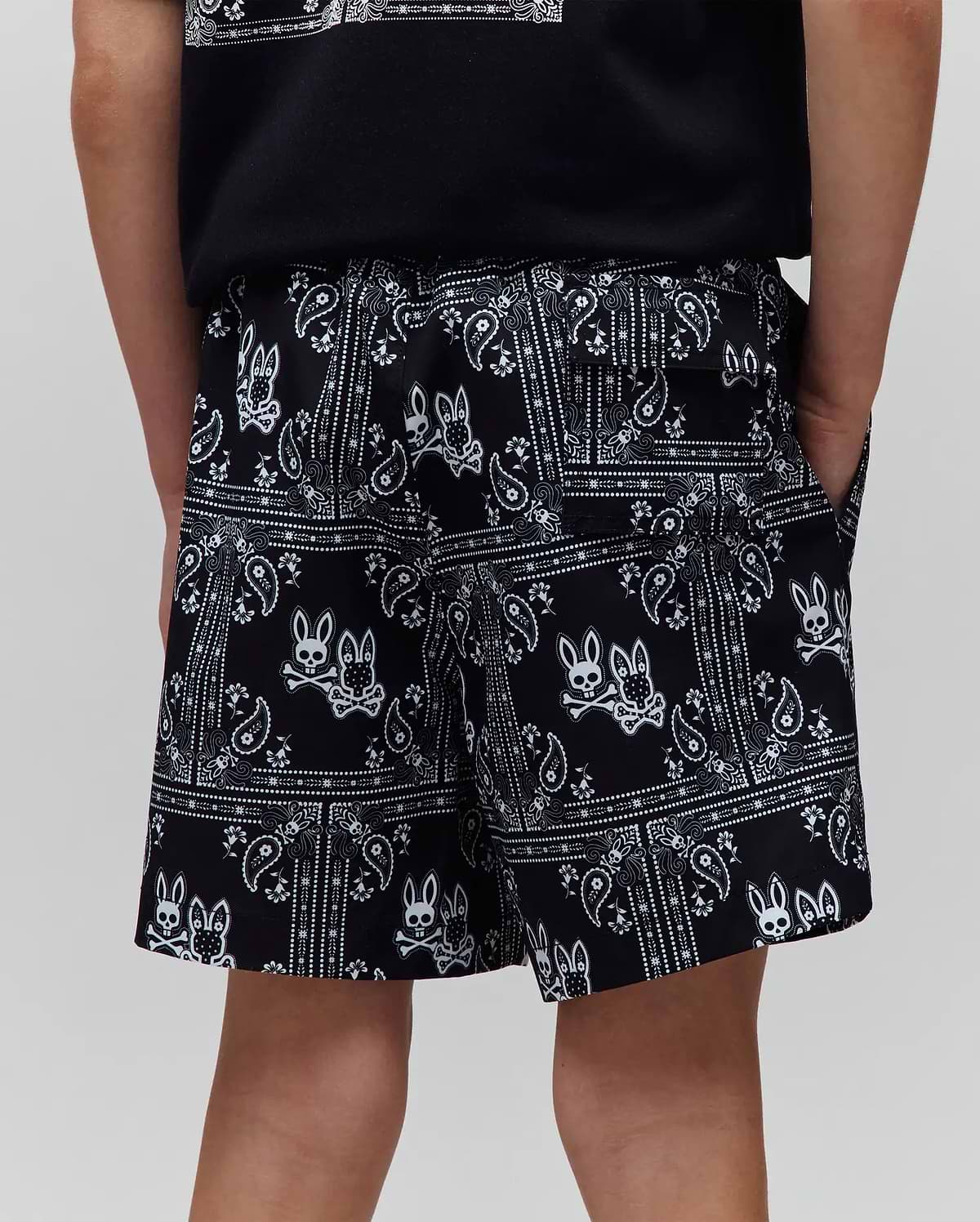 Kids Luke Swim Trunk, black with a vibrant white all-over bandana print, intricately featuring repeated Psycho Bunny skull and crossbones logos, offering a comfortable relaxed fit in a smooth, quick-dry fabric.