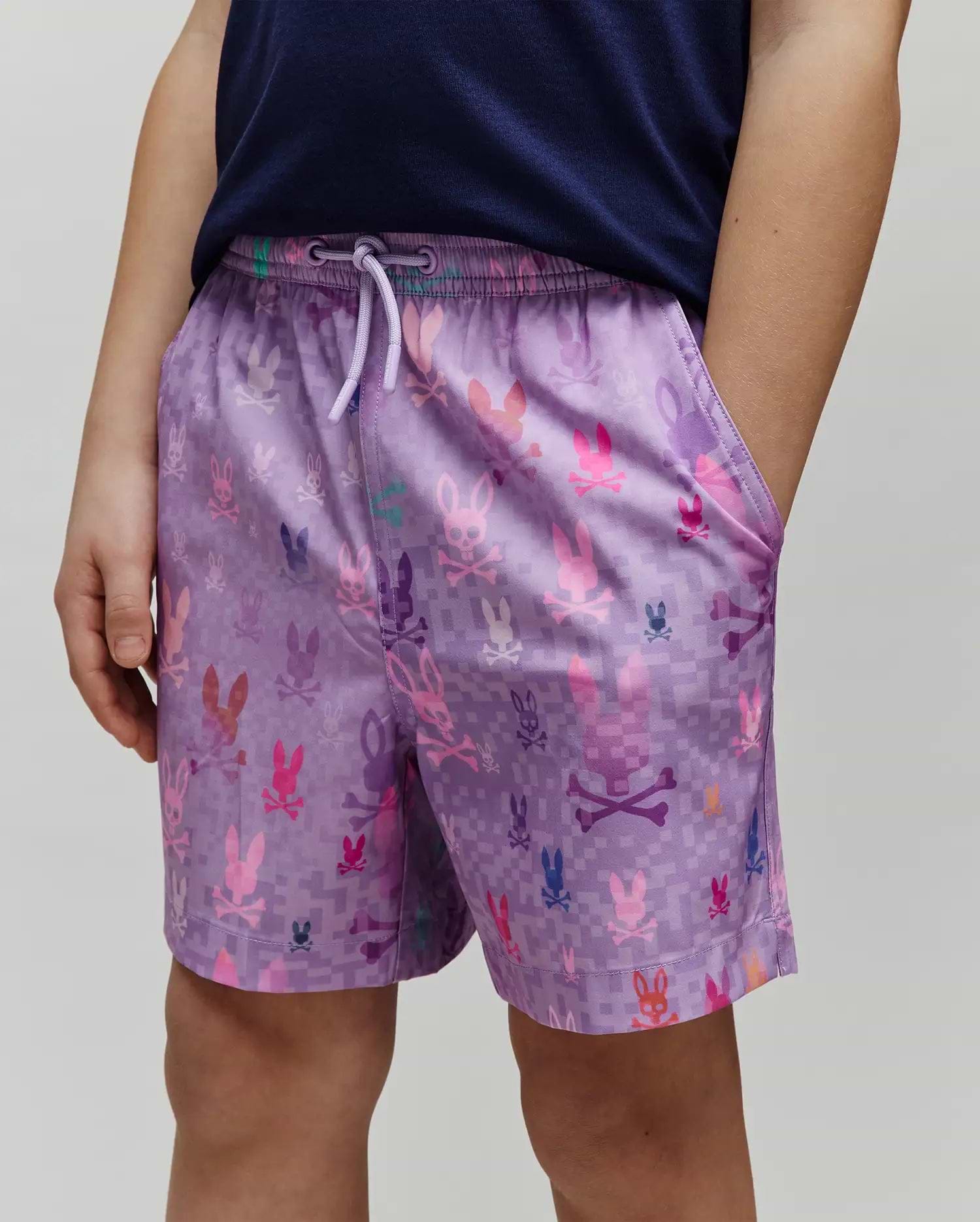 Kids Van Swim Trunk in purple, featuring a relaxed fit and an all-over pixelated print of multi-colored Psycho Bunny skull logos in pink, blue, orange, and green on smooth, quick-dry fabric with a drawstring waist.