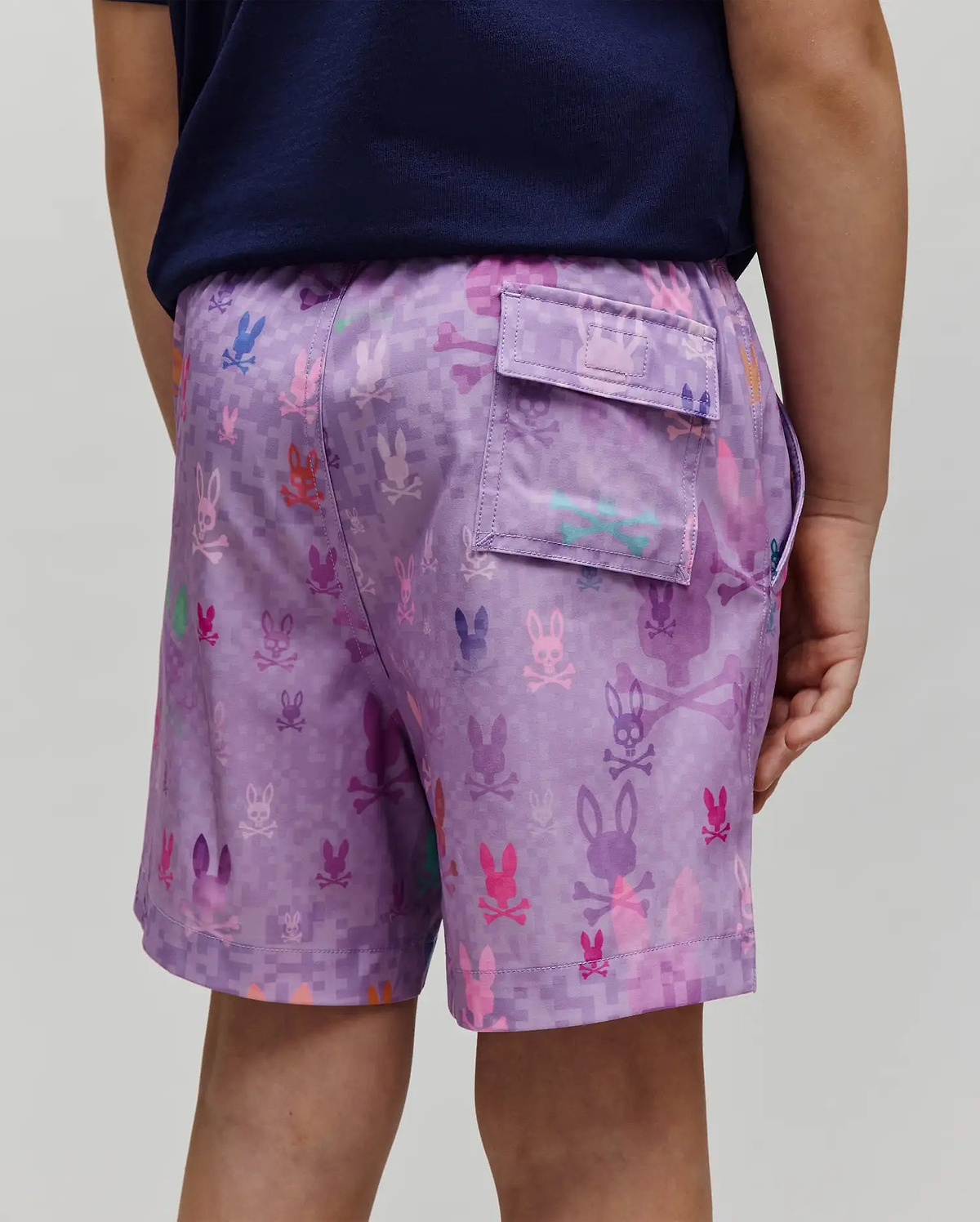Kids Van Swim Trunk in lavender with a pixelated checkerboard pattern and all-over multi-color Psycho Bunny skull logos, featuring a back flap pocket and a relaxed fit in smooth, quick-dry fabric.