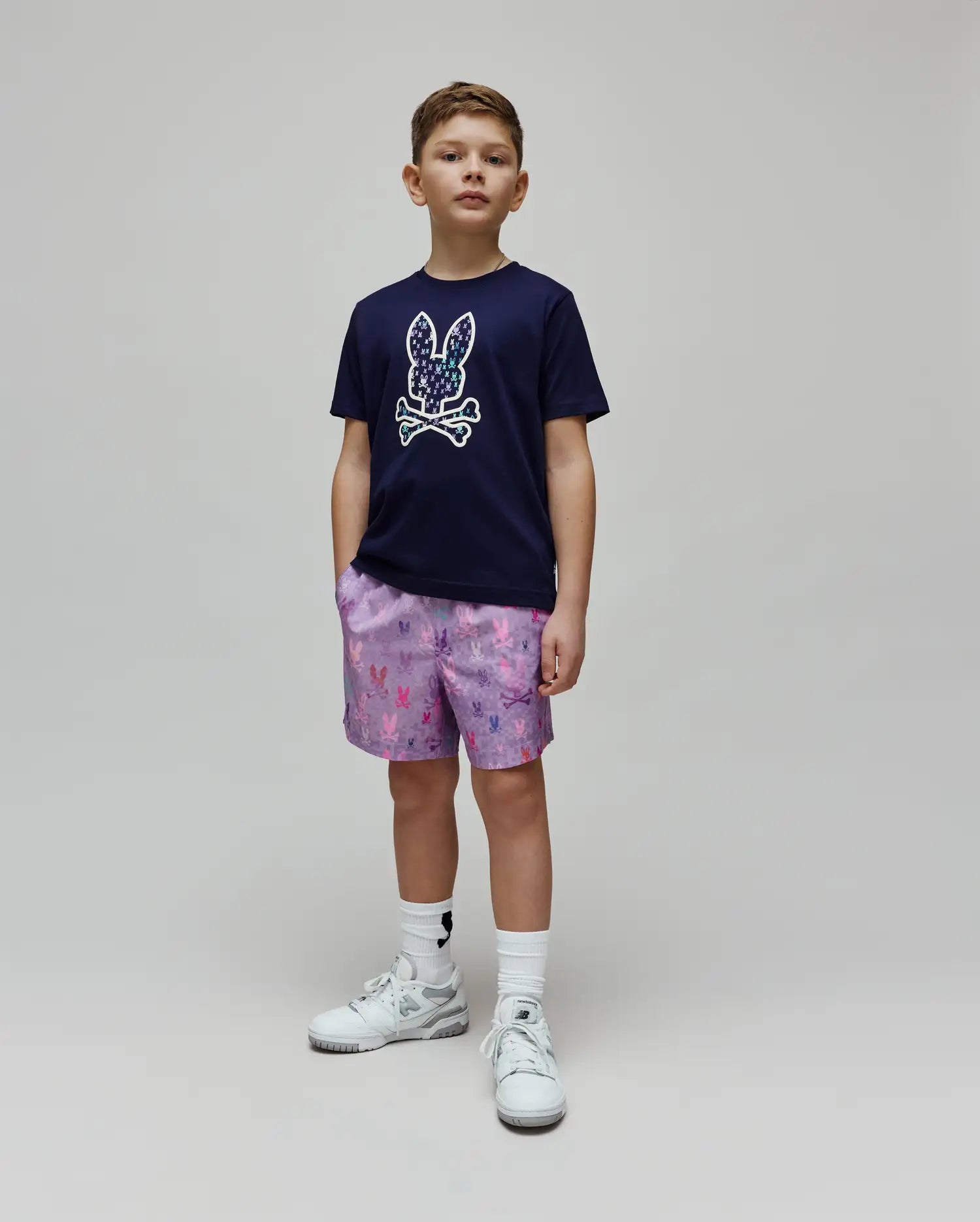 Navy jersey t-shirt with a large chest Psycho Bunny skull logo filled with smaller colorful bunny skulls, paired with lavender Kids Van Swim Trunk featuring an all-over multi-color bunny skull print.