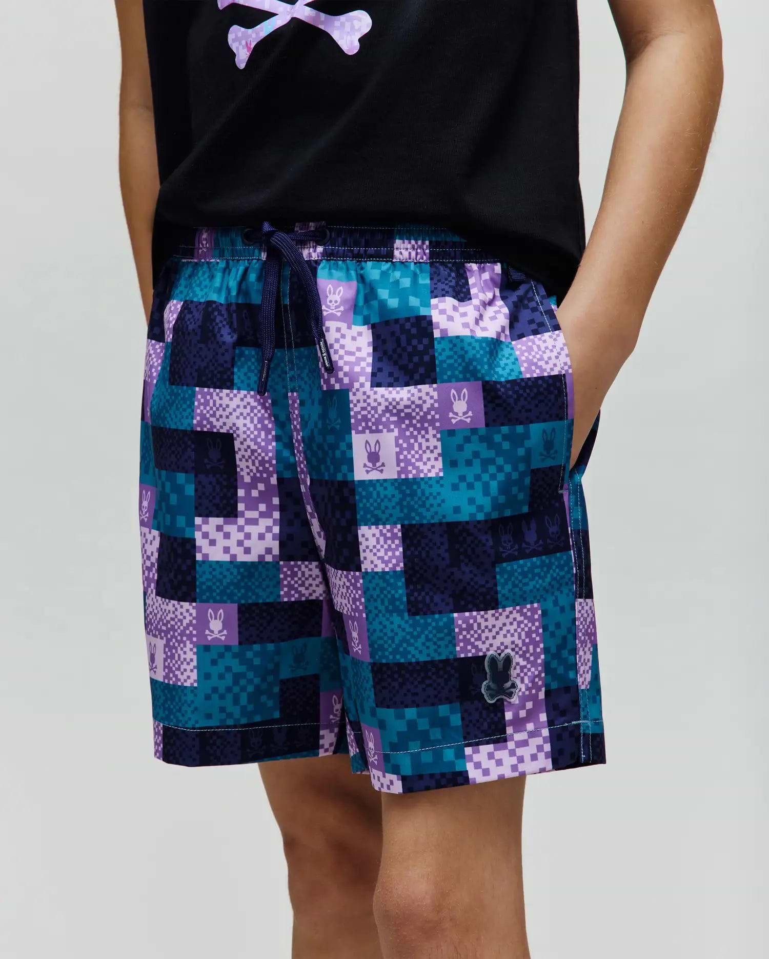 Kids Olivier All Over Bunny Swim Trunk with a vibrant pixelated teal, dark blue, and purple checkered pattern, featuring small bunny skull logos and a rubberized Psycho Bunny logo on the lower left leg.
