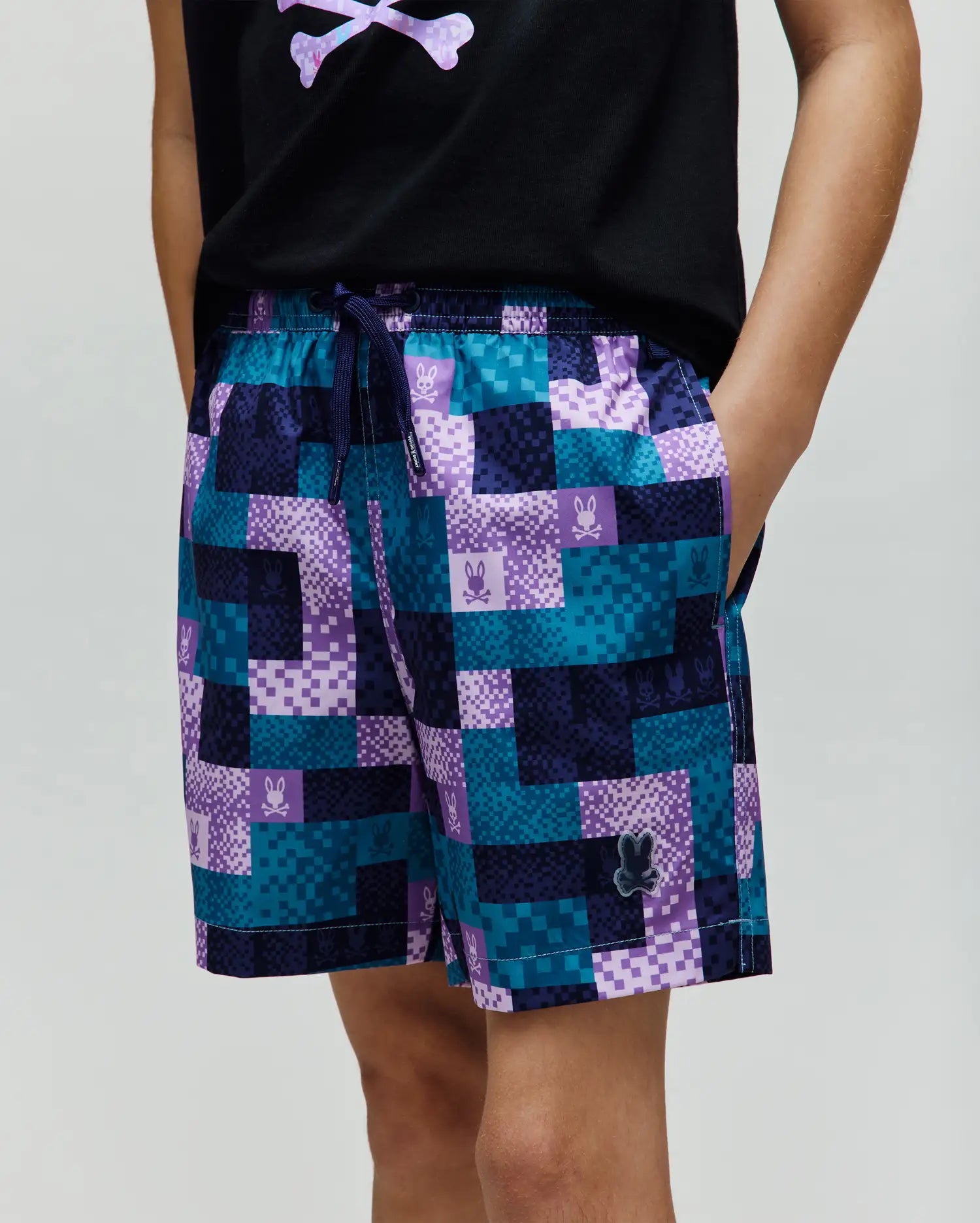 Kids Olivier All Over Bunny Swim Trunk with a vibrant pixelated teal, dark blue, and purple checkered pattern, featuring small bunny skull logos and a rubberized Psycho Bunny logo on the lower left leg.