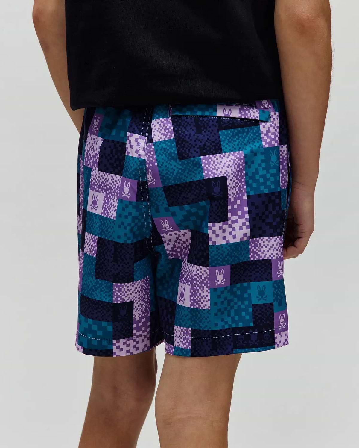 Kids Olivier All Over Bunny Swim Trunk, a relaxed fit swim short with a vibrant pixelated checkerboard pattern in teal, purple, dark blue, and light purple, featuring small repeated Psycho Bunny skull logos throughout the smooth fabric.