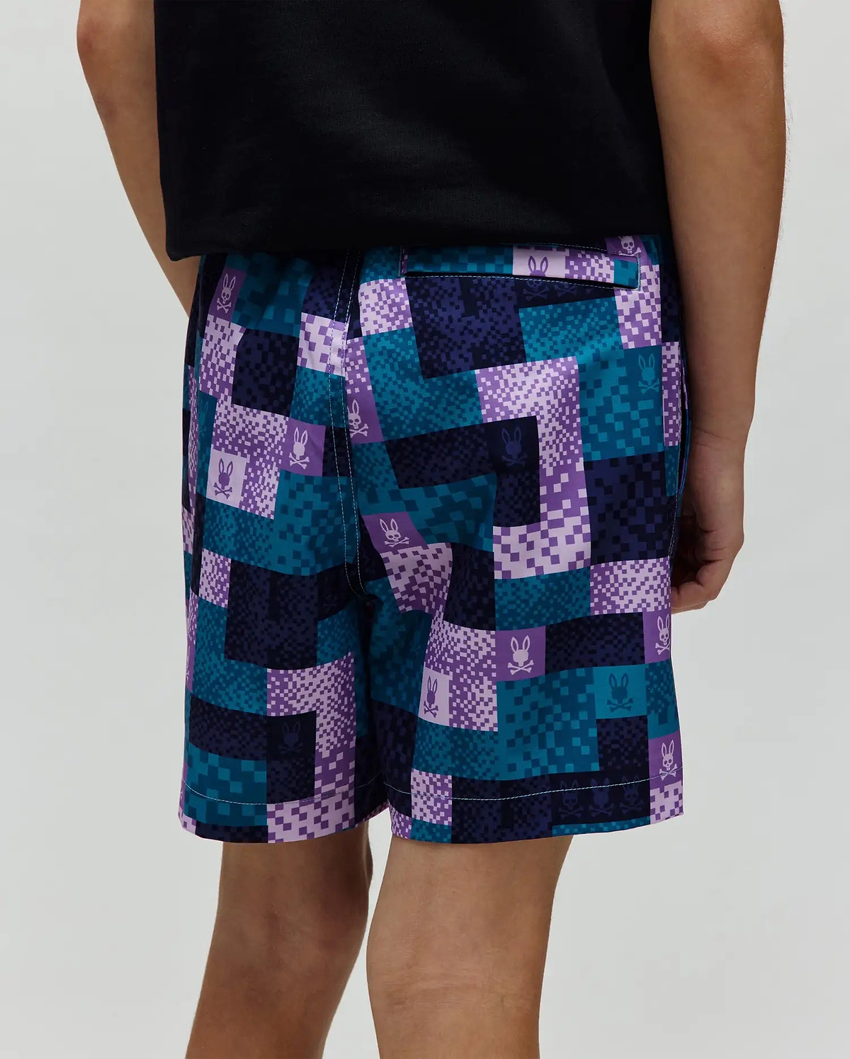 Kids Olivier All Over Bunny Swim Trunk, a relaxed fit swim short with a vibrant pixelated checkerboard pattern in teal, purple, dark blue, and light purple, featuring small repeated Psycho Bunny skull logos throughout the smooth fabric.