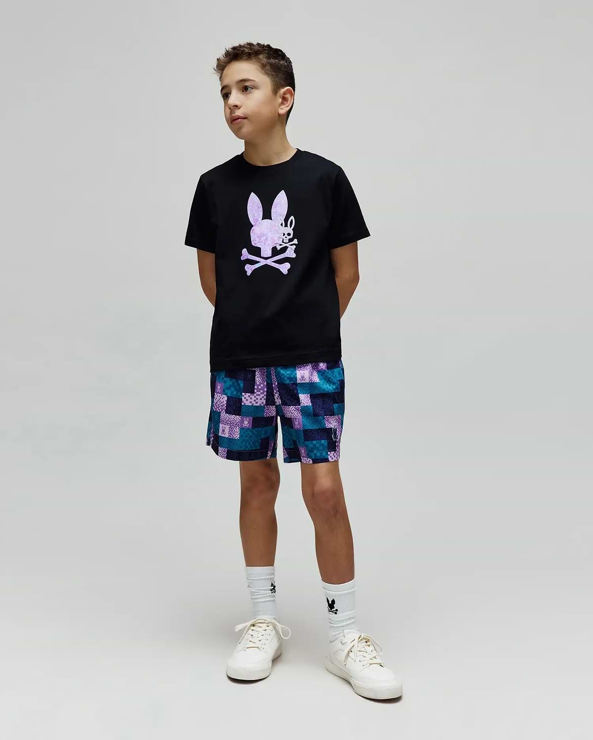 Black classic-fit jersey t-shirt with a large lavender Psycho Bunny skull graphic on the chest, paired with Kids Olivier All Over Bunny Swim Trunk, a blue and purple patchwork short with small bunny logos.