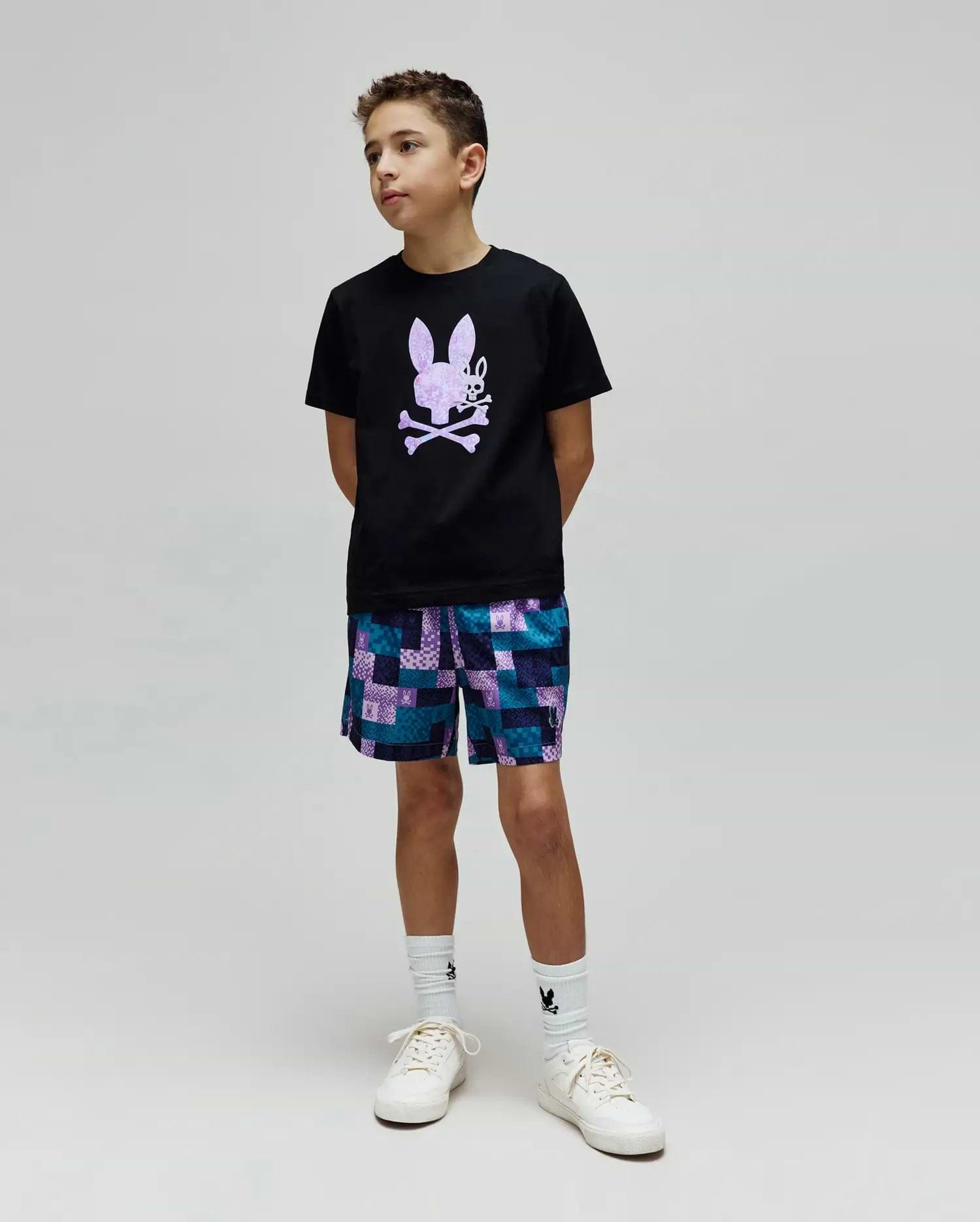 Black classic-fit jersey t-shirt with a large lavender Psycho Bunny skull graphic on the chest, paired with Kids Olivier All Over Bunny Swim Trunk, a blue and purple patchwork short with small bunny logos.