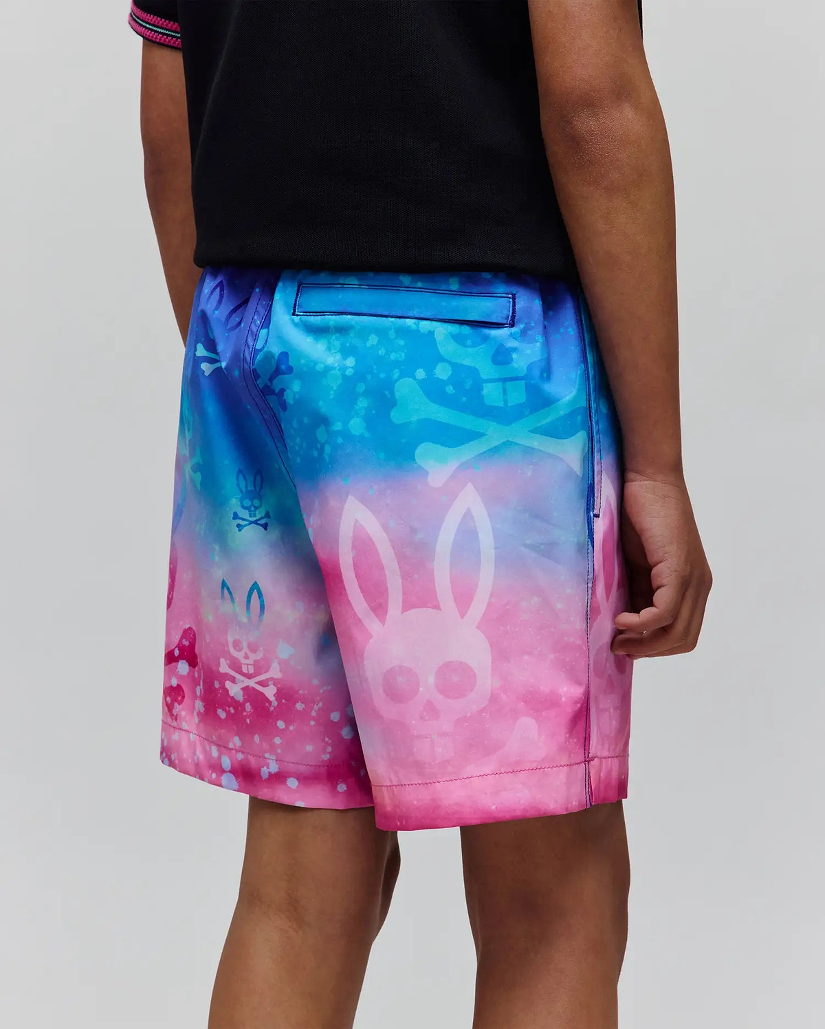Kids Caspian Swim Trunk in a vibrant blue, purple, and pink gradient, featuring an all-over print of Psycho Bunny skull logos and crossbones on smooth, quick-dry fabric with a relaxed fit.