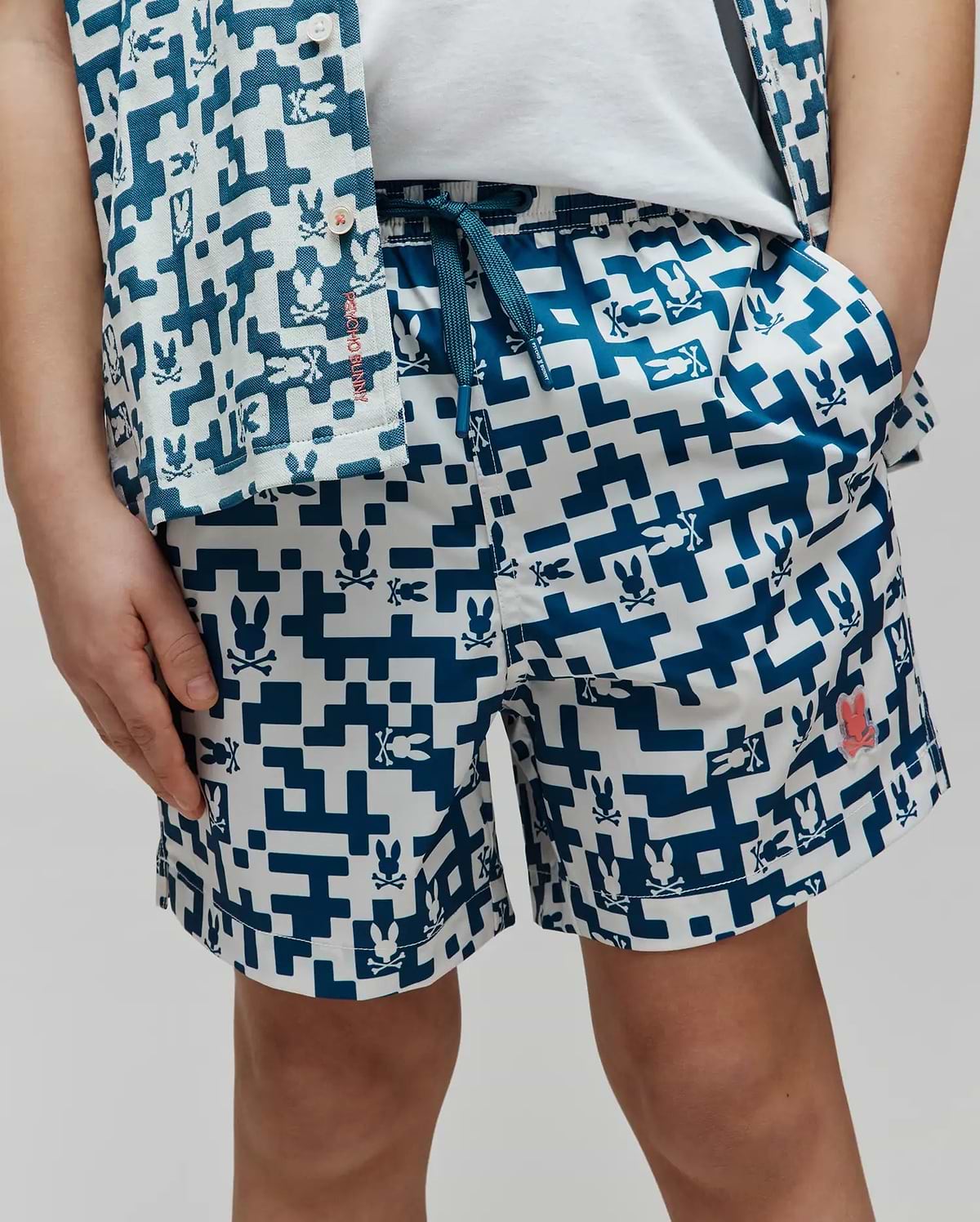 Kids Josh Swim Trunk in a relaxed fit, featuring a white and teal pixelated geometric pattern with integrated Psycho Bunny skull logos, smooth quick-dry fabric, a teal drawstring, and a small coral logo patch on the left leg. (2)