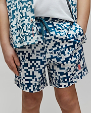 Kids Josh Swim Trunk in a relaxed fit, featuring a white and teal pixelated geometric pattern with integrated Psycho Bunny skull logos, smooth quick-dry fabric, a teal drawstring, and a small coral logo patch on the left leg. (2)