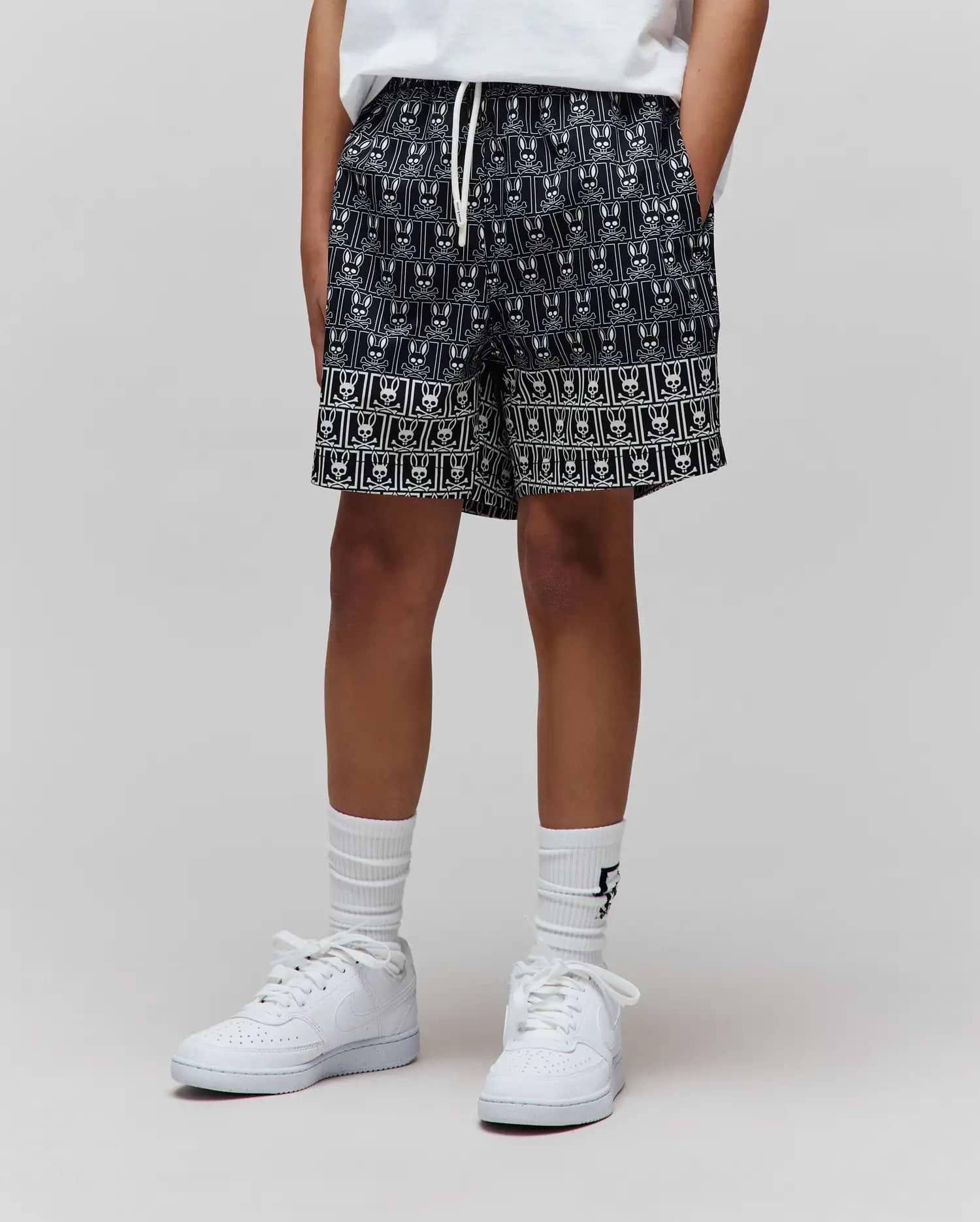 Kids Keon Swim Trunk, black with an all-over white Psycho Bunny skull logo pattern, featuring a distinct border pattern at the hem, relaxed fit, smooth woven fabric, and a white drawstring. (1)