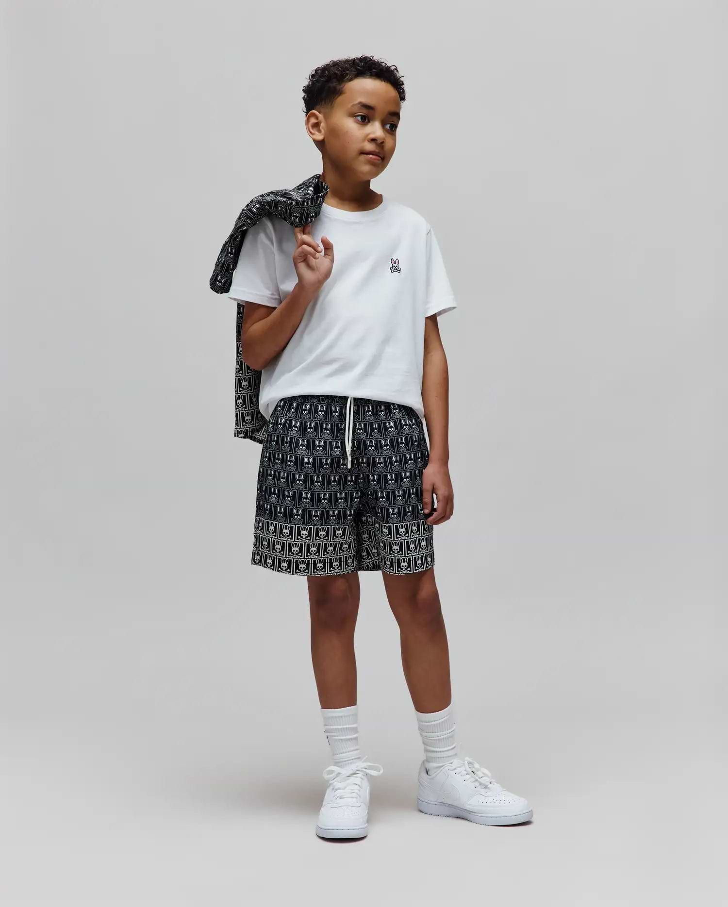 White classic fit jersey t-shirt with small embroidered Psycho Bunny logo on chest. Paired with Kids Keon Swim Trunk, black with all-over white bunny skull print, and a matching patterned jacket. (4)