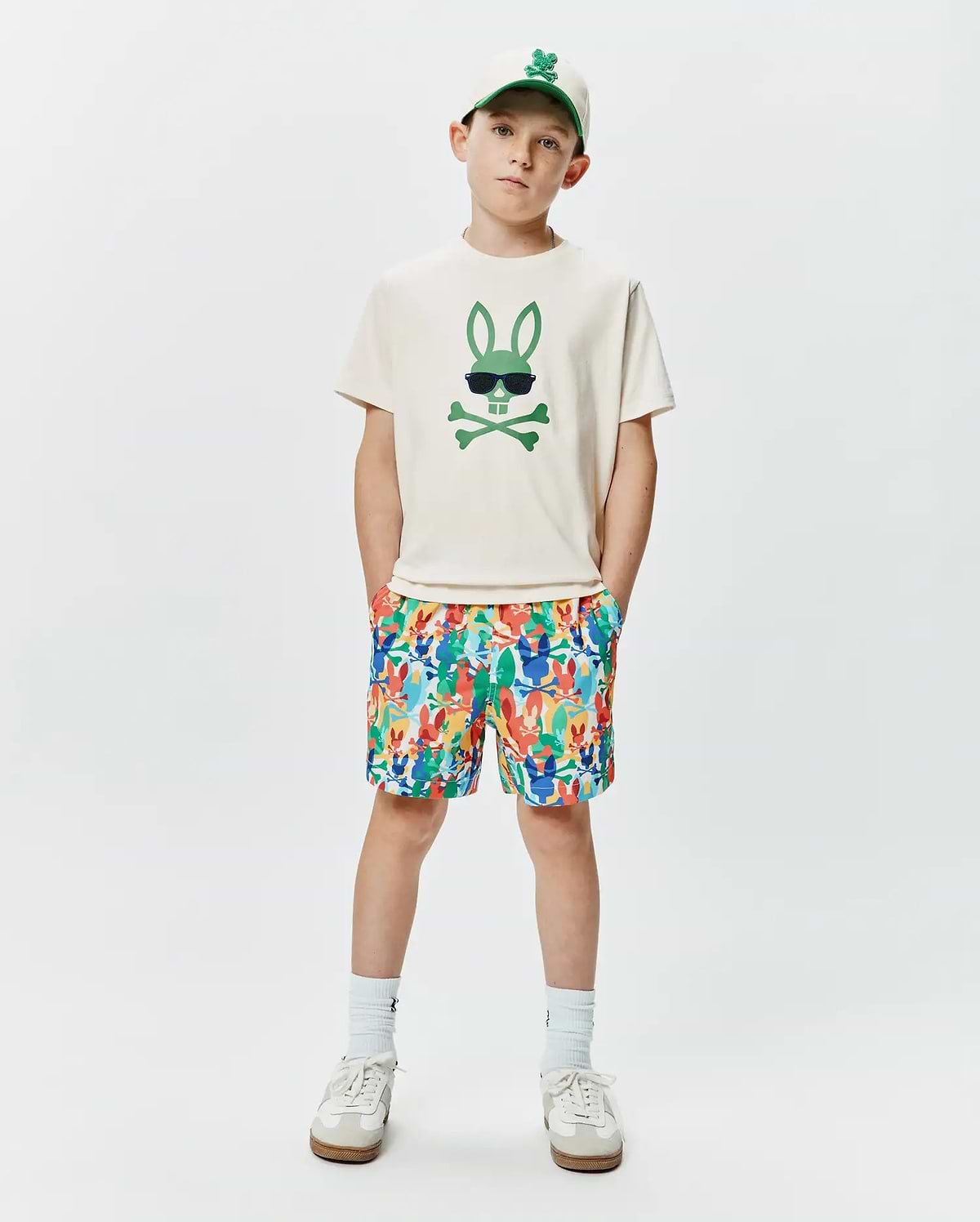 Off-white jersey t-shirt with a large green Psycho Bunny skull graphic, paired with multicolor KIDS TANNER SWIM TRUNK featuring an all-over bunny skull print, and a cream baseball cap with an embroidered green bunny logo.