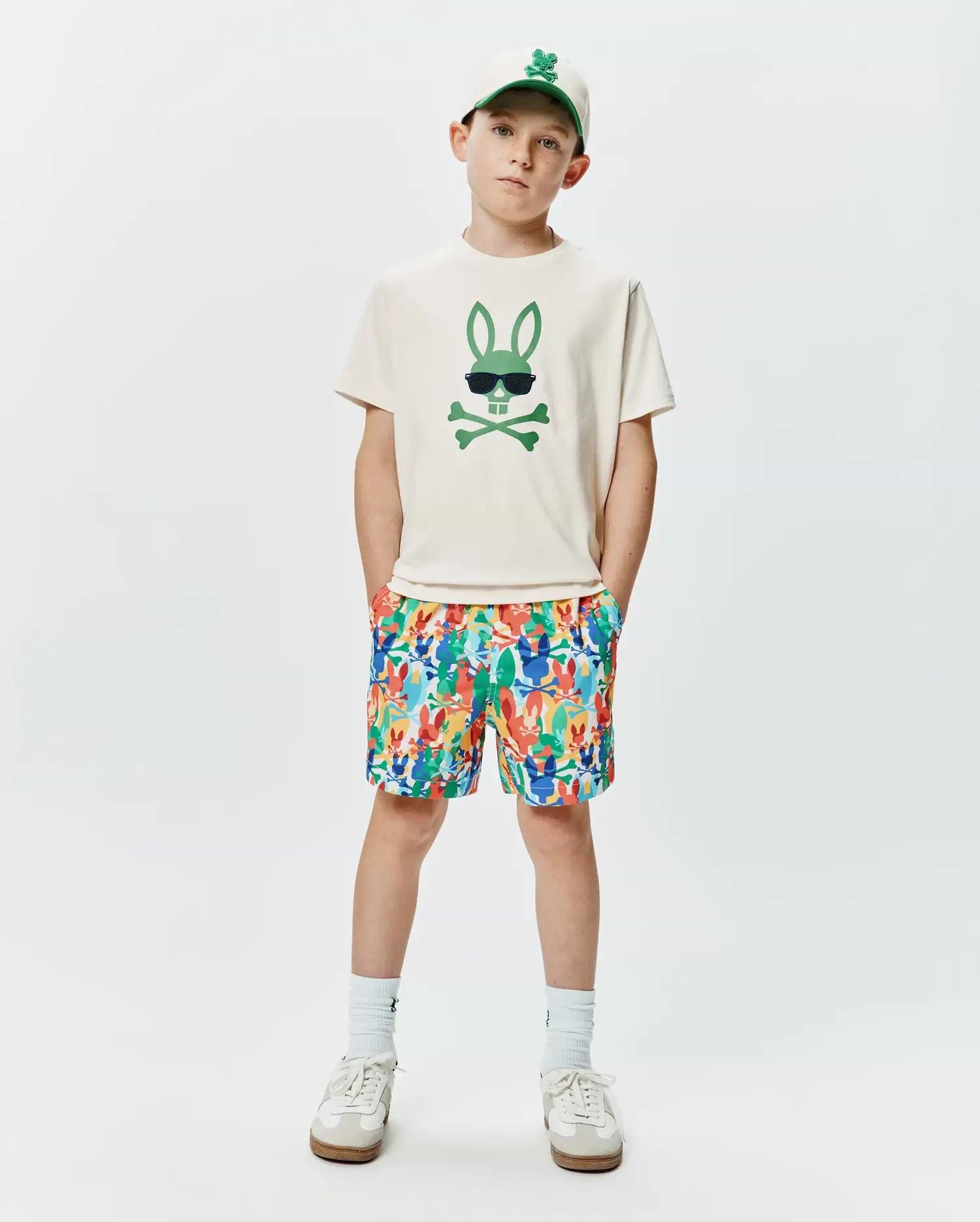 Off-white jersey t-shirt with a large green Psycho Bunny skull graphic, paired with multicolor KIDS TANNER SWIM TRUNK featuring an all-over bunny skull print, and a cream baseball cap with an embroidered green bunny logo.