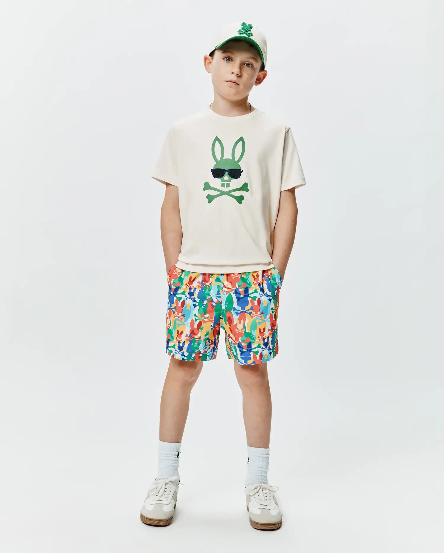 Off-white jersey t-shirt with a large green Psycho Bunny skull graphic, paired with multicolor KIDS TANNER SWIM TRUNK featuring an all-over bunny skull print, and a cream baseball cap with an embroidered green bunny logo.