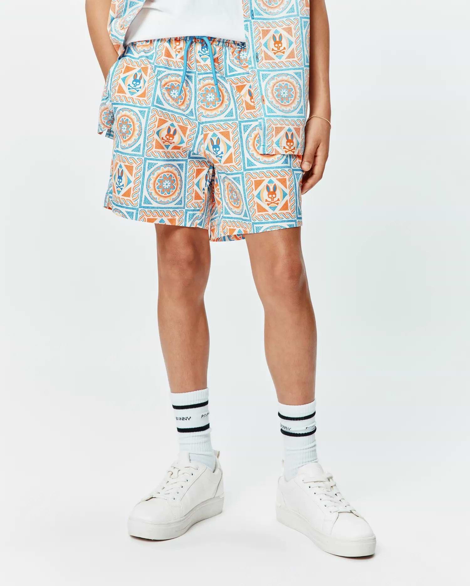 Kids Frank Swim Trunk in white with a vibrant blue and orange mosaic pattern, featuring repeated Psycho Bunny skull logos and a bright blue drawstring, in a relaxed fit. (1)
