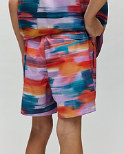 Kids Raphael Swim Trunk, a Psycho Bunny piece, showcasing a vibrant multi-color brushstroke pattern in teal, orange, red, purple, and pink, with a relaxed fit and smooth fabric.