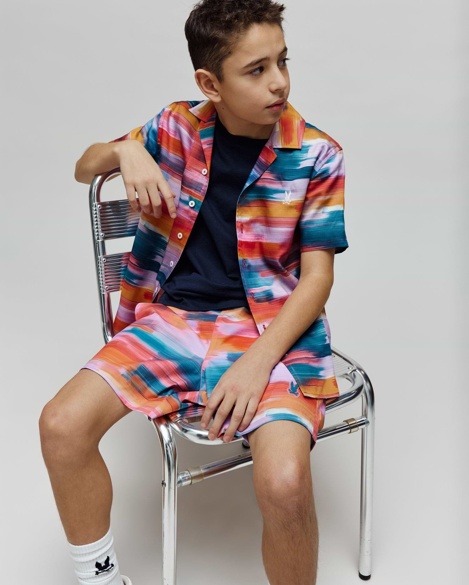 Vibrant multi-color brushstroke short-sleeve button-up shirt and matching Kids Raphael Swim Trunk, both featuring the small embroidered Psycho Bunny logo. A navy crew neck t-shirt is worn underneath.