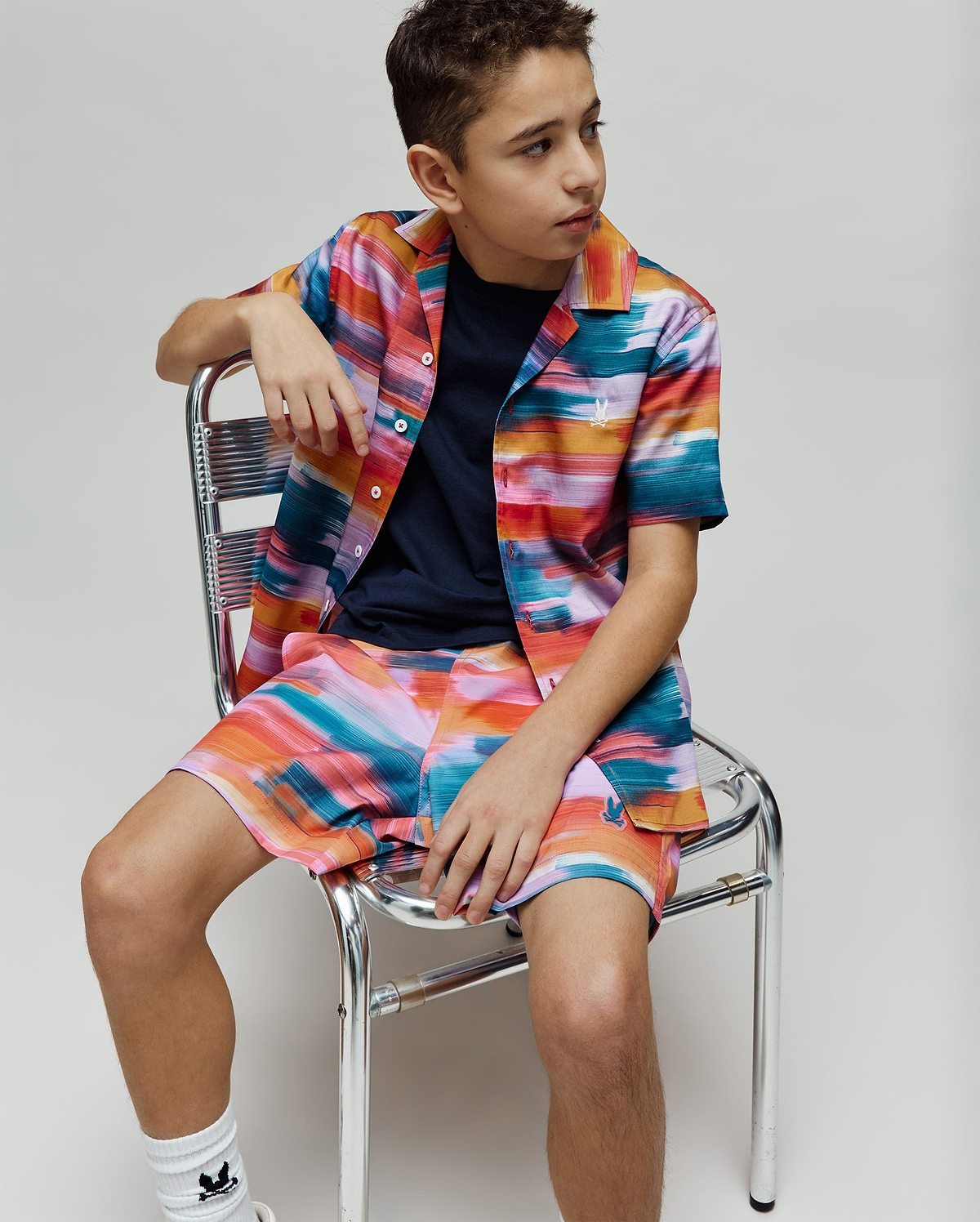 Vibrant multi-color brushstroke short-sleeve button-up shirt and matching Kids Raphael Swim Trunk, both featuring the small embroidered Psycho Bunny logo. A navy crew neck t-shirt is worn underneath.