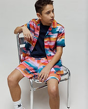 Vibrant multi-color brushstroke short-sleeve button-up shirt and matching Kids Raphael Swim Trunk, both featuring the small embroidered Psycho Bunny logo. A navy crew neck t-shirt is worn underneath.