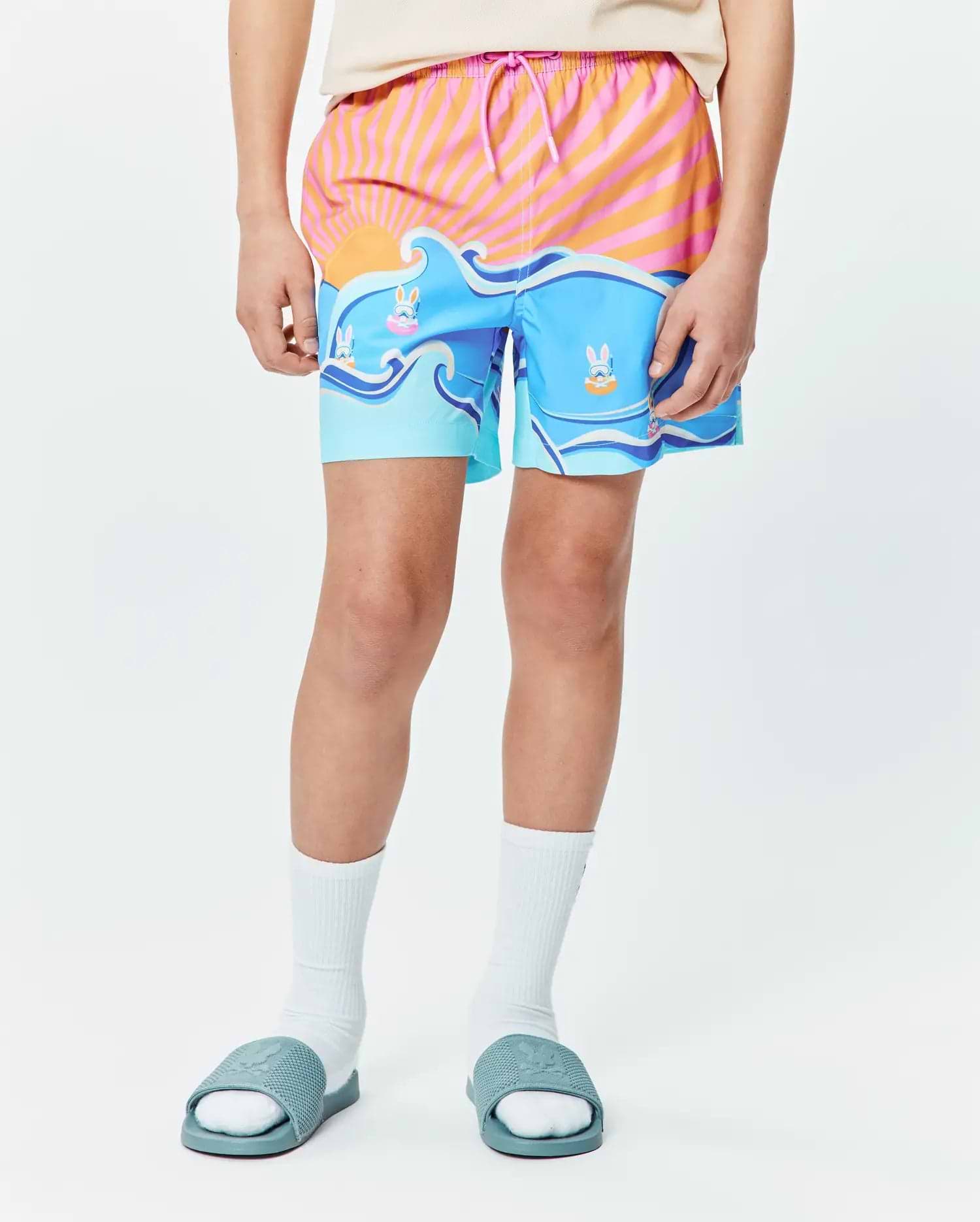 Kids Ray Swim Trunk 6 with a vibrant graphic print featuring an orange and pink striped sunrise over blue ocean waves, accented by playful Psycho Bunny skull logos in goggles floating on inner tubes.