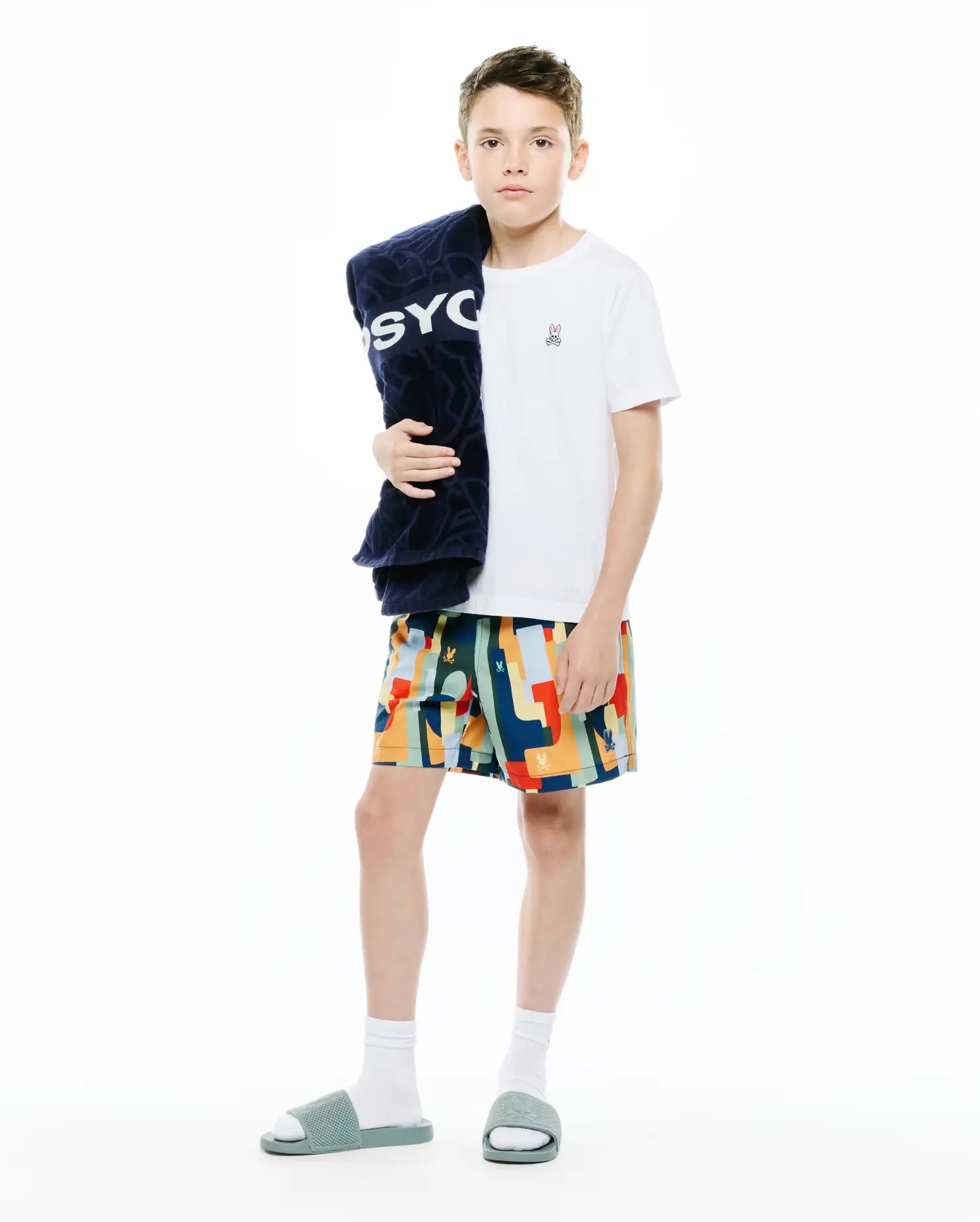 White jersey t-shirt with small embroidered Psycho Bunny logo, paired with Kids Wes All Over Print Swim Trunk featuring a vibrant geometric pattern and logo, plus a navy terry towel with large PSYCHO text.