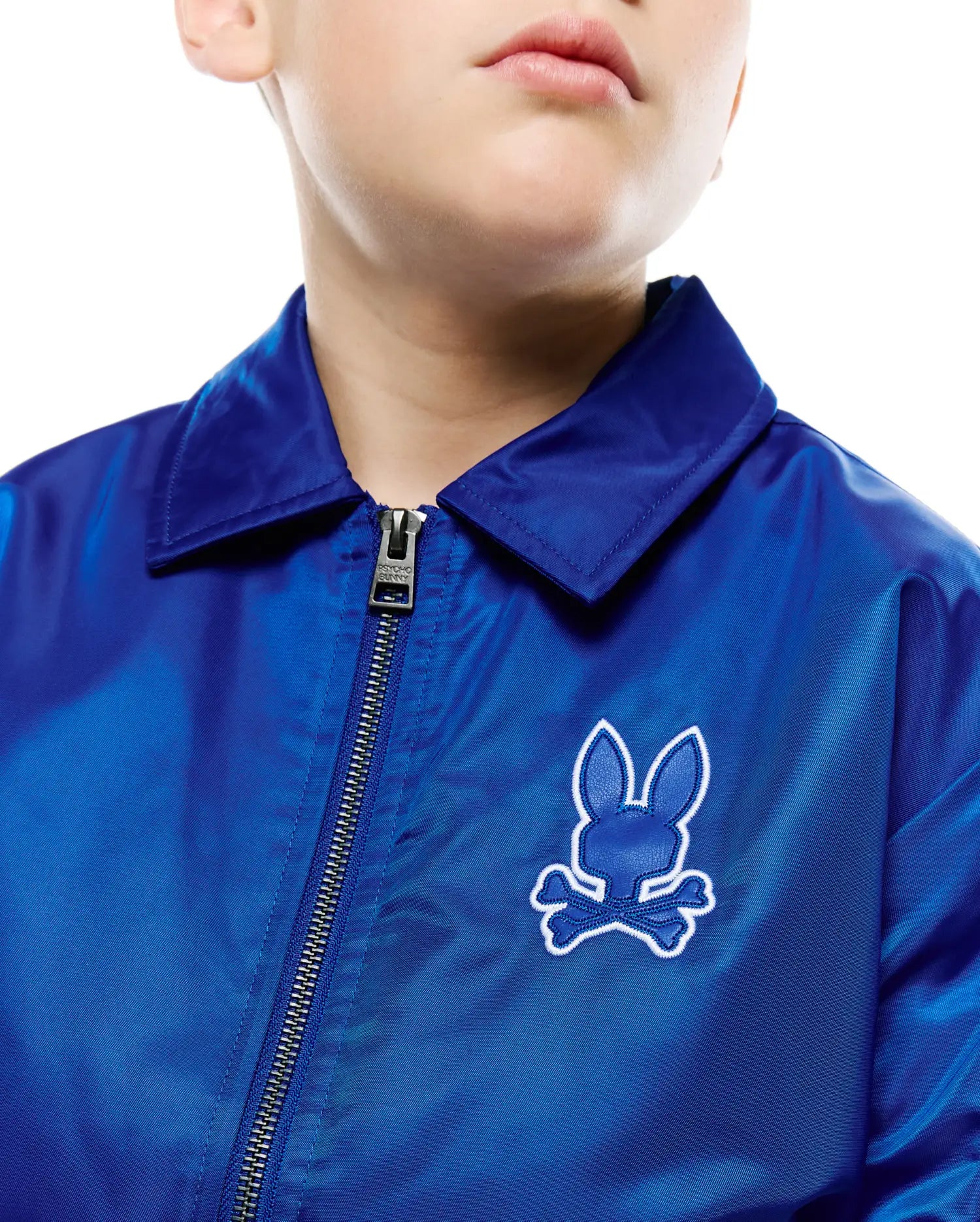 Kids Nathaniel Satin Coach Jacket in royal blue satin, featuring a collared neck, full silver zipper with Psycho Bunny pull, and an embroidered royal blue bunny skull logo with white outline on the left chest.