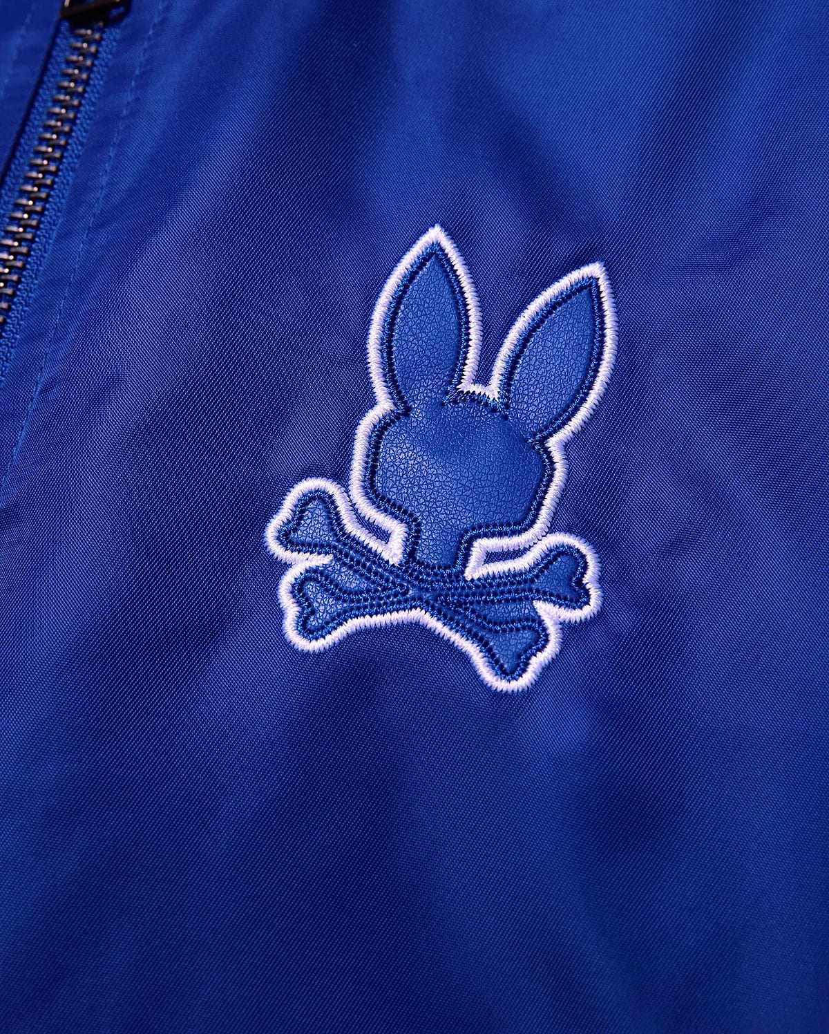 Kids Nathaniel Satin Coach Jacket in smooth royal blue, featuring a prominent textured dark blue Psycho Bunny skull logo with a thick white embroidered outline.