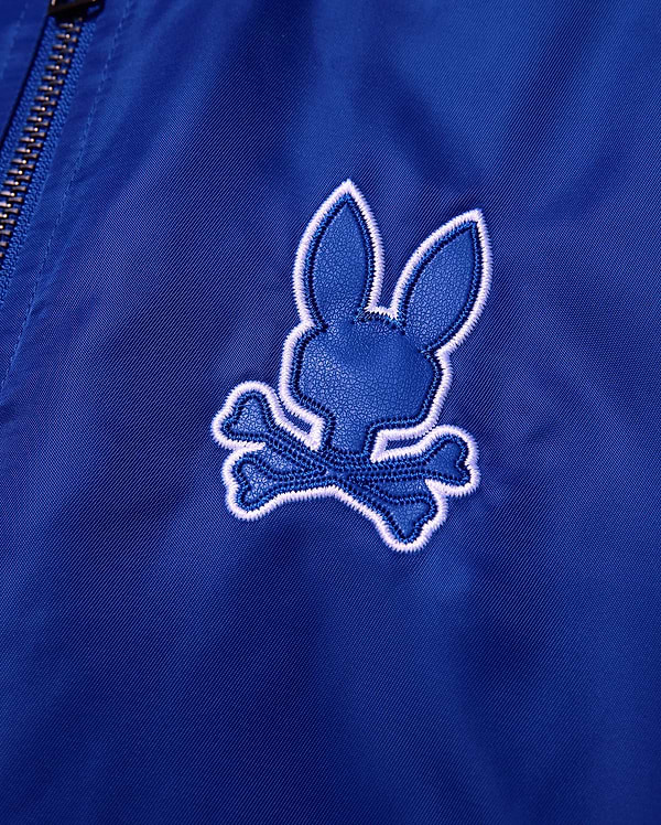 Kids Nathaniel Satin Coach Jacket in smooth royal blue, featuring a prominent textured dark blue Psycho Bunny skull logo with a thick white embroidered outline.