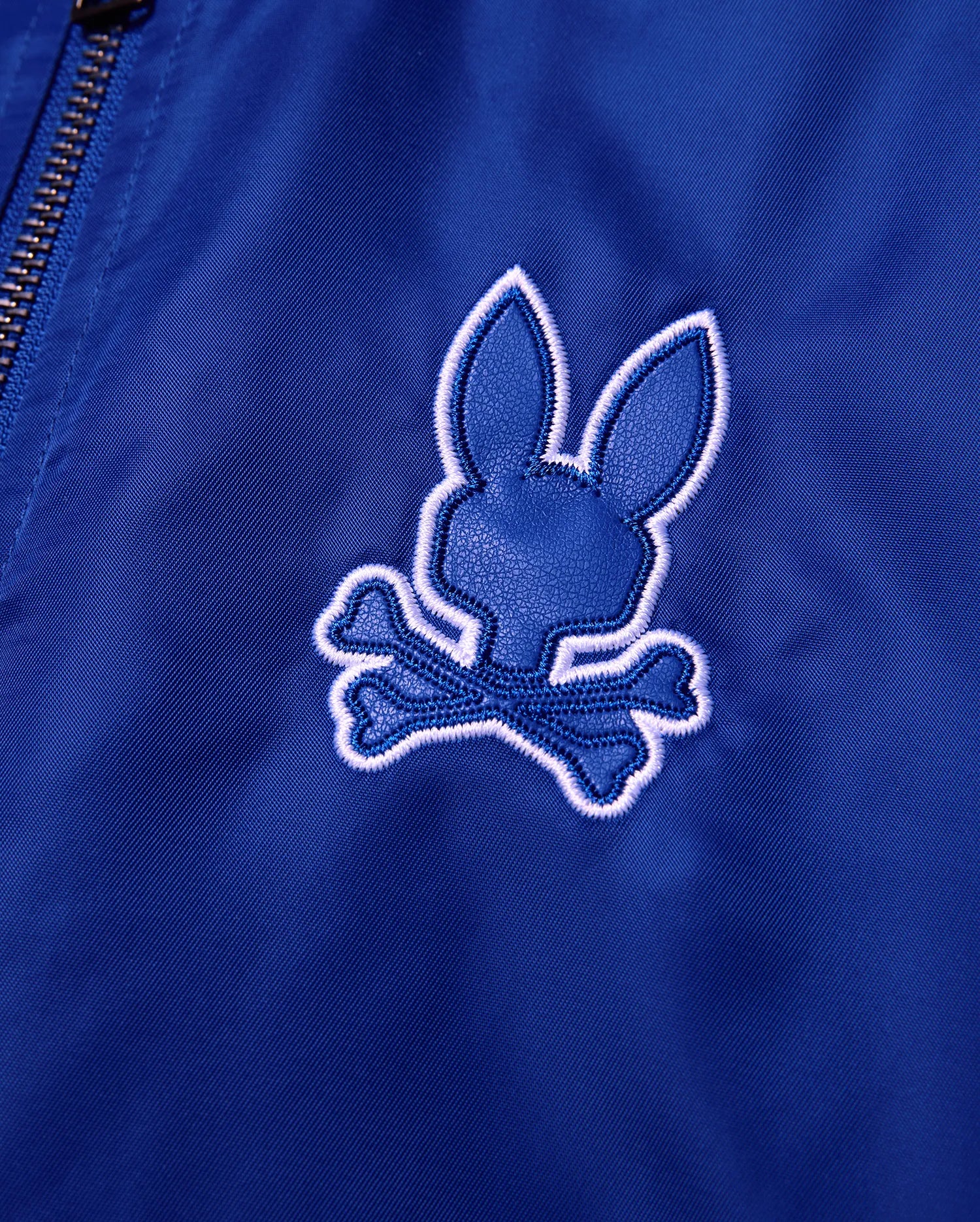 Kids Nathaniel Satin Coach Jacket in smooth royal blue, featuring a prominent textured dark blue Psycho Bunny skull logo with a thick white embroidered outline.