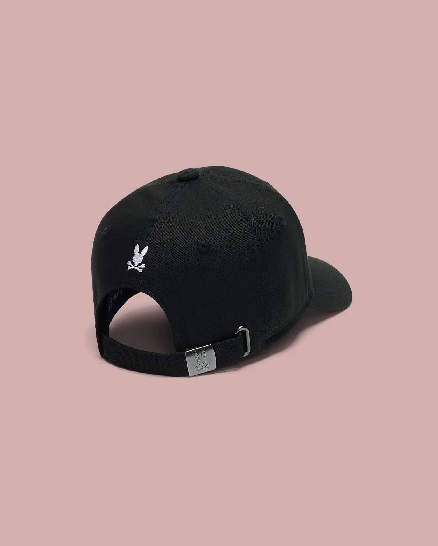 The Psycho Bunny KIDS ARCHIVE BASEBALL CAP (KO1500004) in black cotton twill is shown from the back, featuring a small embroidered Bunny logo above the opening and an adjustable strap with metal clasp against a pink background.
