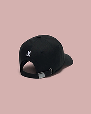 The Psycho Bunny KIDS ARCHIVE BASEBALL CAP (KO1500004) in black cotton twill is shown from the back, featuring a small embroidered Bunny logo above the opening and an adjustable strap with metal clasp against a pink background.