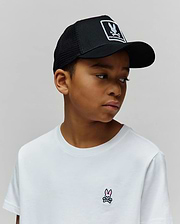 White classic fit jersey t-shirt with small pink/black embroidered Psycho Bunny skull logo on chest, and a black mesh KIDS ADAMO TRUCKER CAP with large white Psycho Bunny skull logo patch.