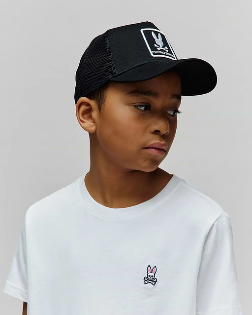 White classic fit jersey t-shirt with small pink/black embroidered Psycho Bunny skull logo on chest, and a black mesh KIDS ADAMO TRUCKER CAP with large white Psycho Bunny skull logo patch.