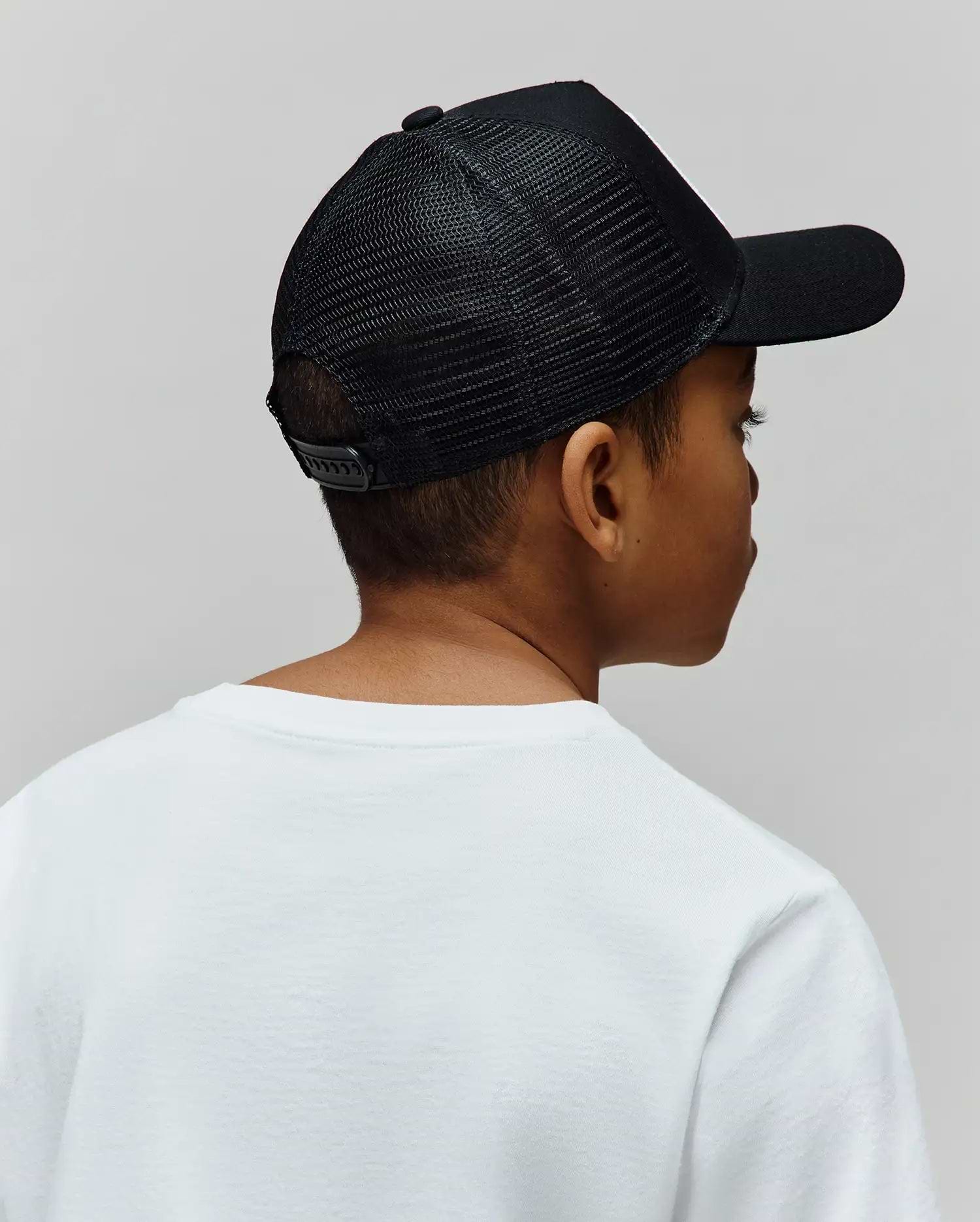 Black mesh back Kids Adamo Trucker Cap featuring a structured solid front panel, curved brim, and adjustable snapback closure, worn with a classic fit plain white jersey knit crew neck t-shirt.
