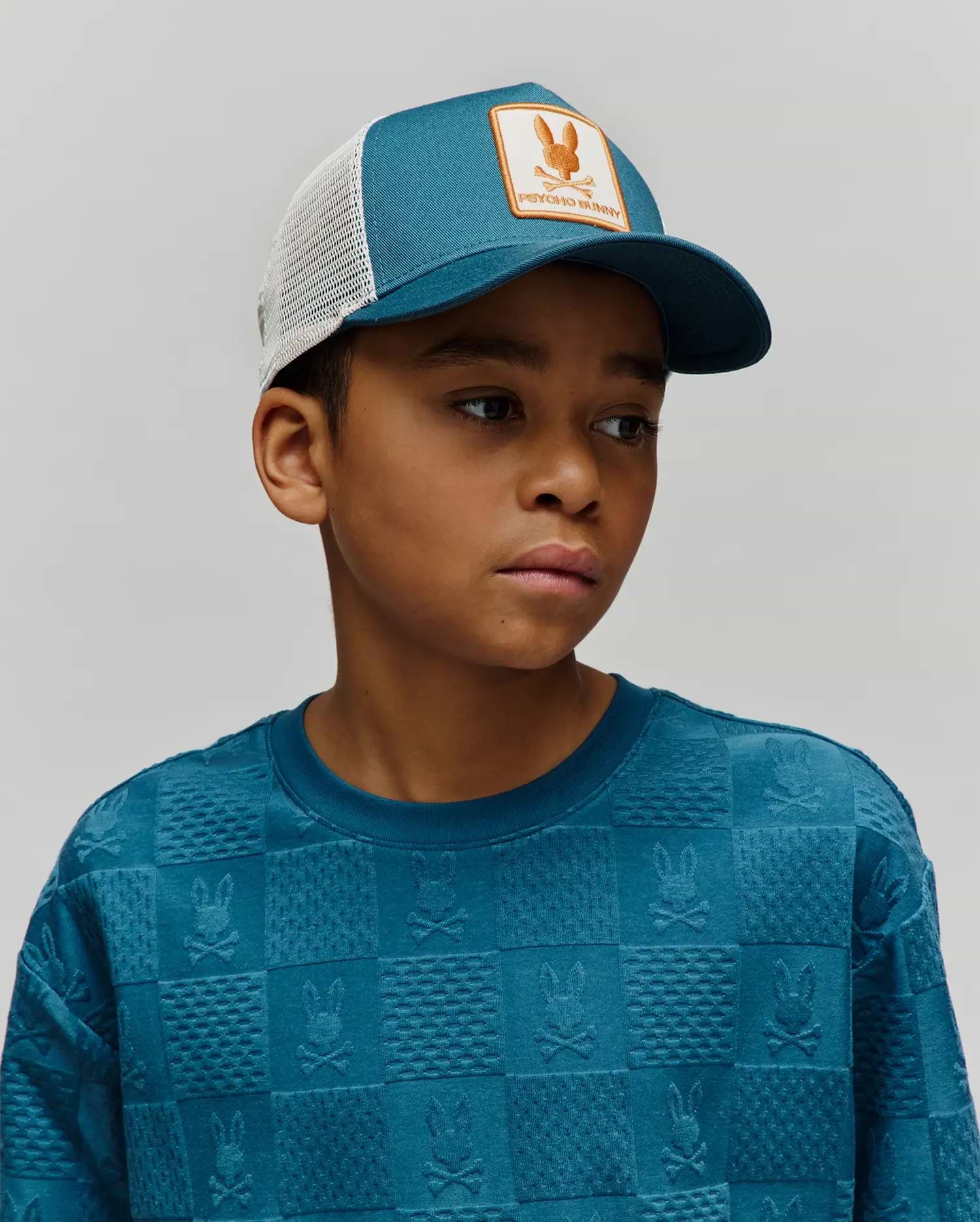 Kids Adamo Trucker Cap, teal front, white mesh, orange Psycho Bunny skull logo patch, and a teal crewneck sweatshirt with an embossed checkerboard Psycho Bunny skull pattern.